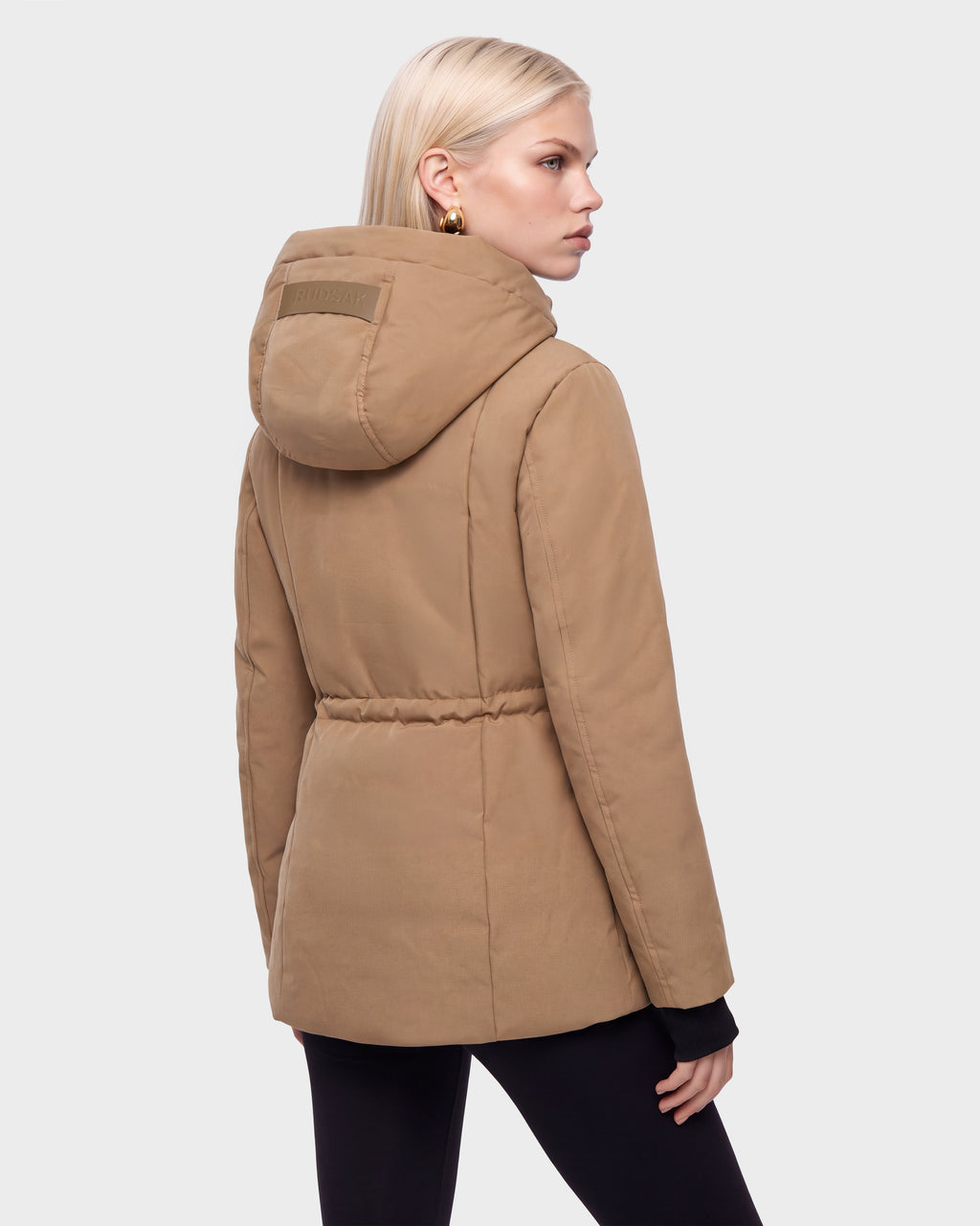 A woman with blond hair models the Rudsak OPAL - 8126618 Camel parka, standing against a light gray background, facing away and looking over her left shoulder in black leggings.