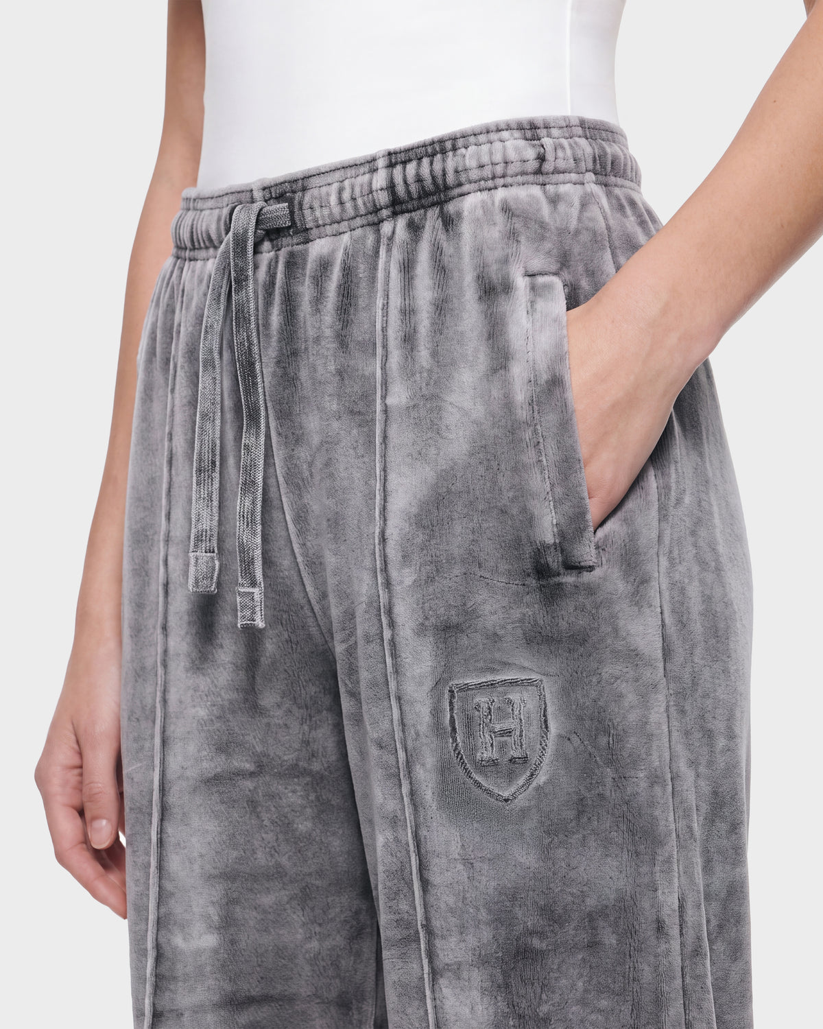 A person is wearing the Rudsak OLIVYA HV - H526903 Titanium gray velour sweatpants, which have a drawstring waist, a pocket with their hand inside, and an embroidered crest logo on the left leg. They are also wearing a white top.