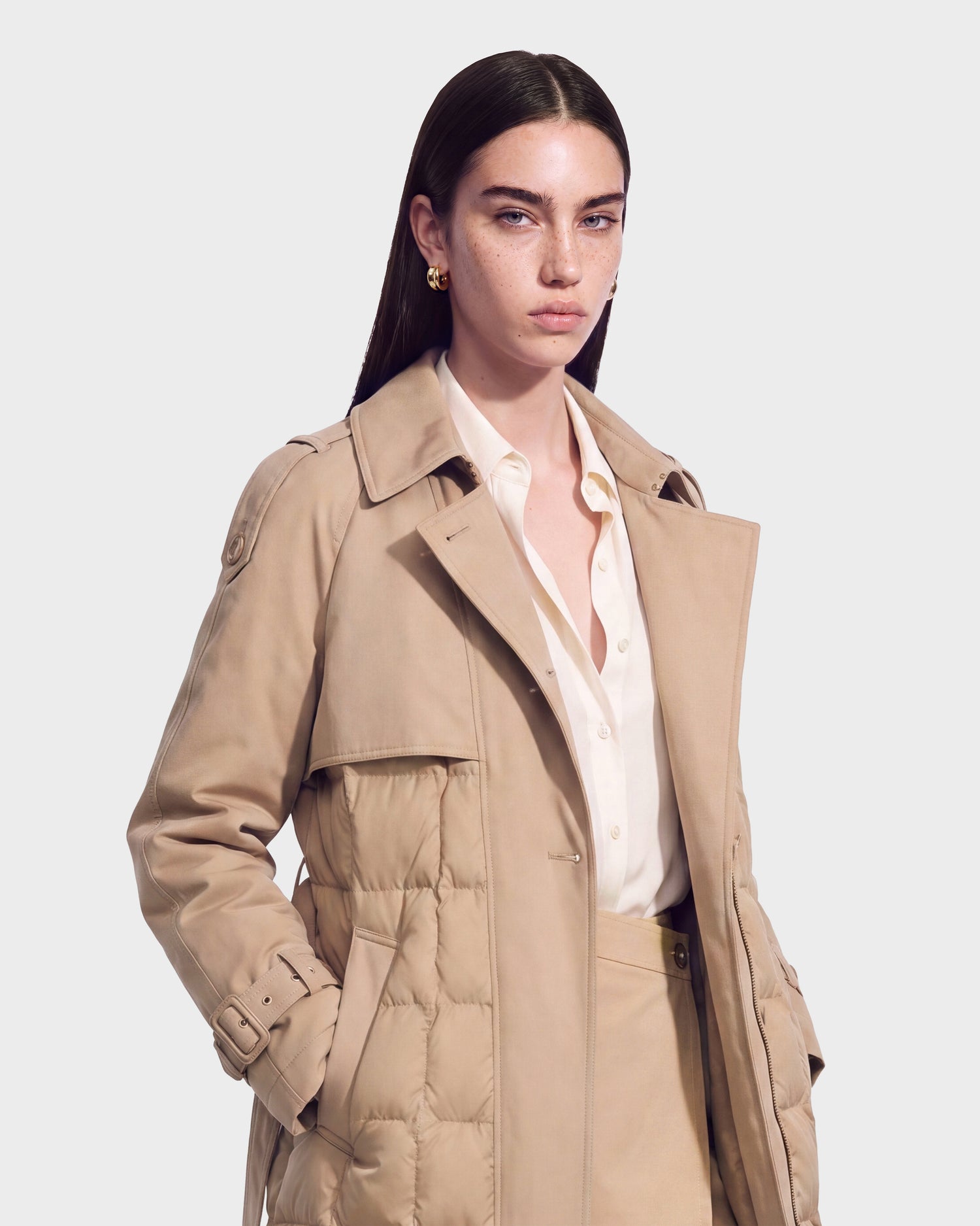 A woman with straight brown hair wears the Rudsak ELVIE - 8126512 Almond beige down trench coat over a cream blouse and matching pants. She stands against a plain light background, looking forward with a neutral expression.