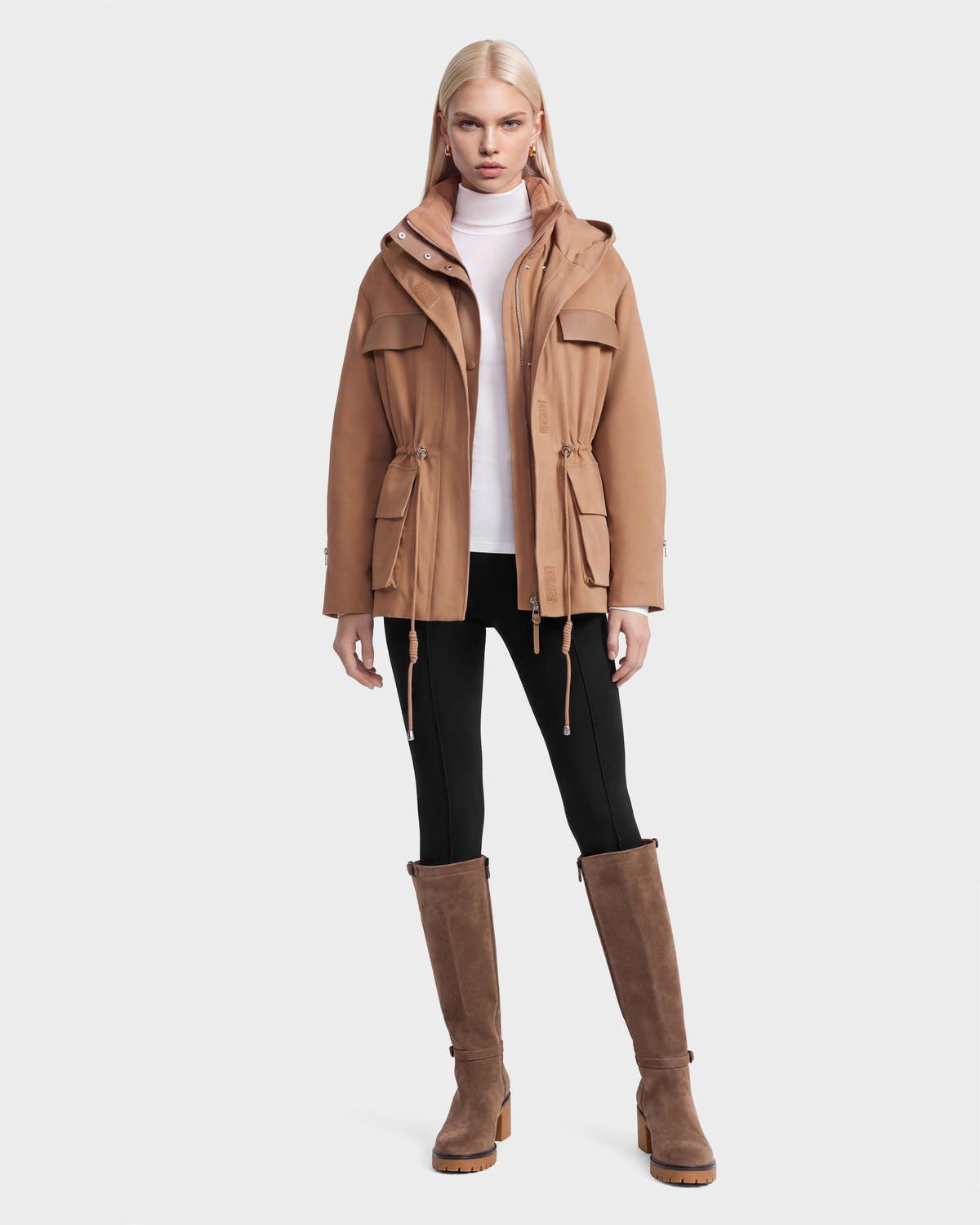 A woman wears a camel-colored Rudsak ROSALINDA - 8126523 hooded jacket with a drawstring waist, a white turtleneck, black pants, and knee-high brown boots.