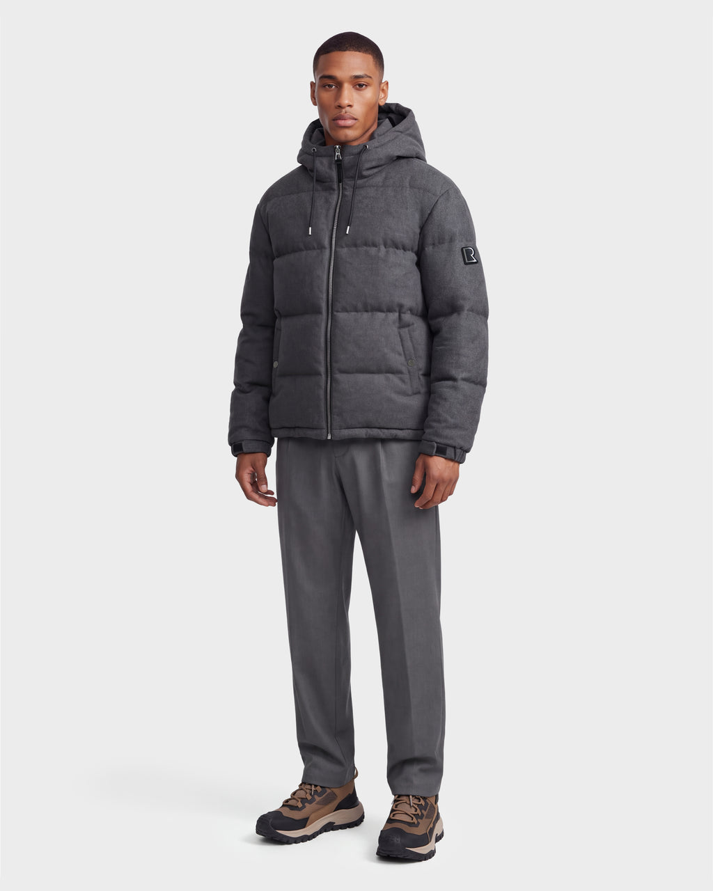 A man stands against a plain white background wearing the Rudsak STERLING W - 6126760 Heather Charcoal puffer jacket with a hood, gray trousers, and brown-and-black sneakers.