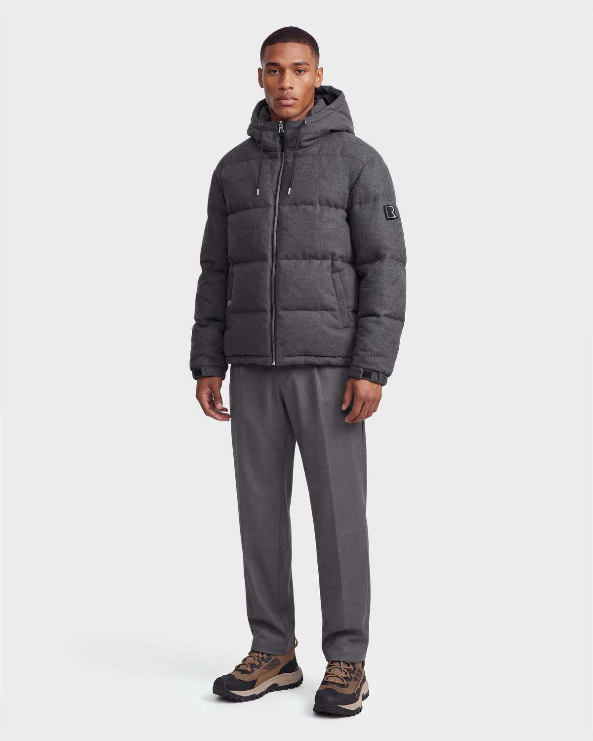 A man stands against a plain white background wearing the Rudsak STERLING W - 6126760 Heather Charcoal puffer jacket with a hood, gray trousers, and brown-and-black sneakers.