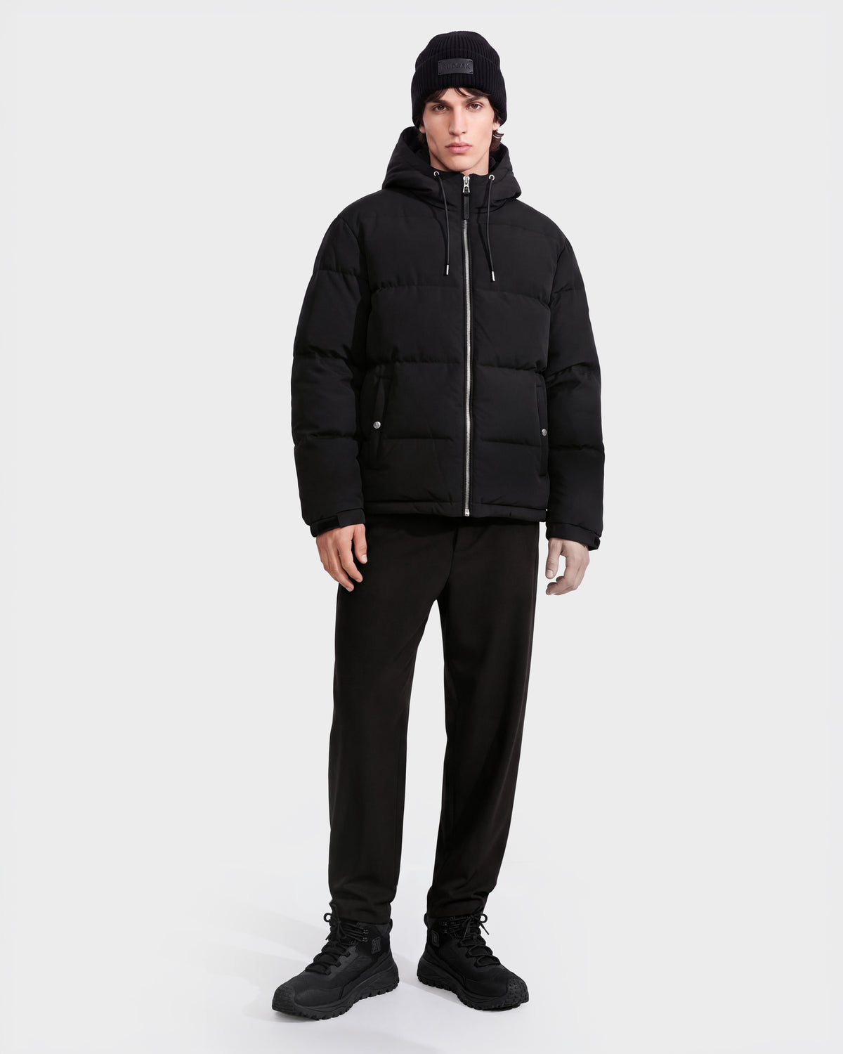 A person stands facing forward against a plain white background, wearing the Rudsak STERLING EL - 6126759 Black puffer jacket, paired with a black beanie, black pants, and black sneakers.