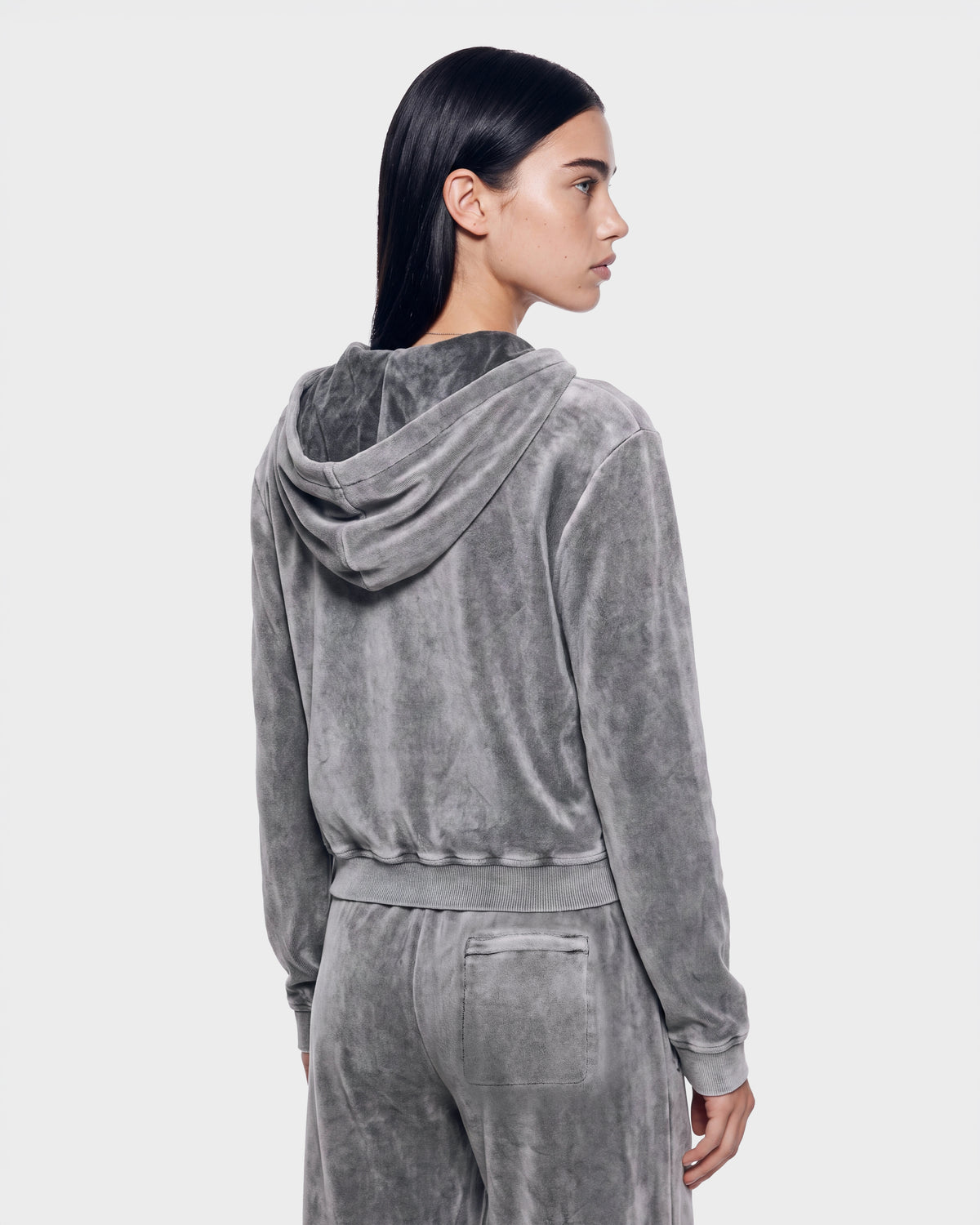 A woman with straight dark hair models the Rudsak TYRA HV - H526904 Titanium velour hoodie and matching pants. She is facing away, turning slightly to the side to showcase the back and side profile of the outfit against a light gray background.