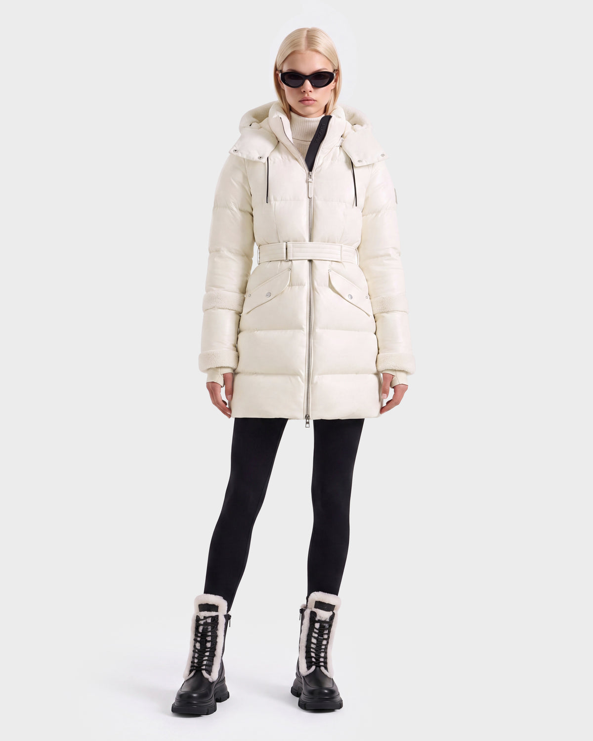 A woman wears a vanilla-colored AUDREY L - 8124585 knee-length down puffer jacket belted over a turtleneck, black leggings, and black lace-up boots with fur-trimmed collars.