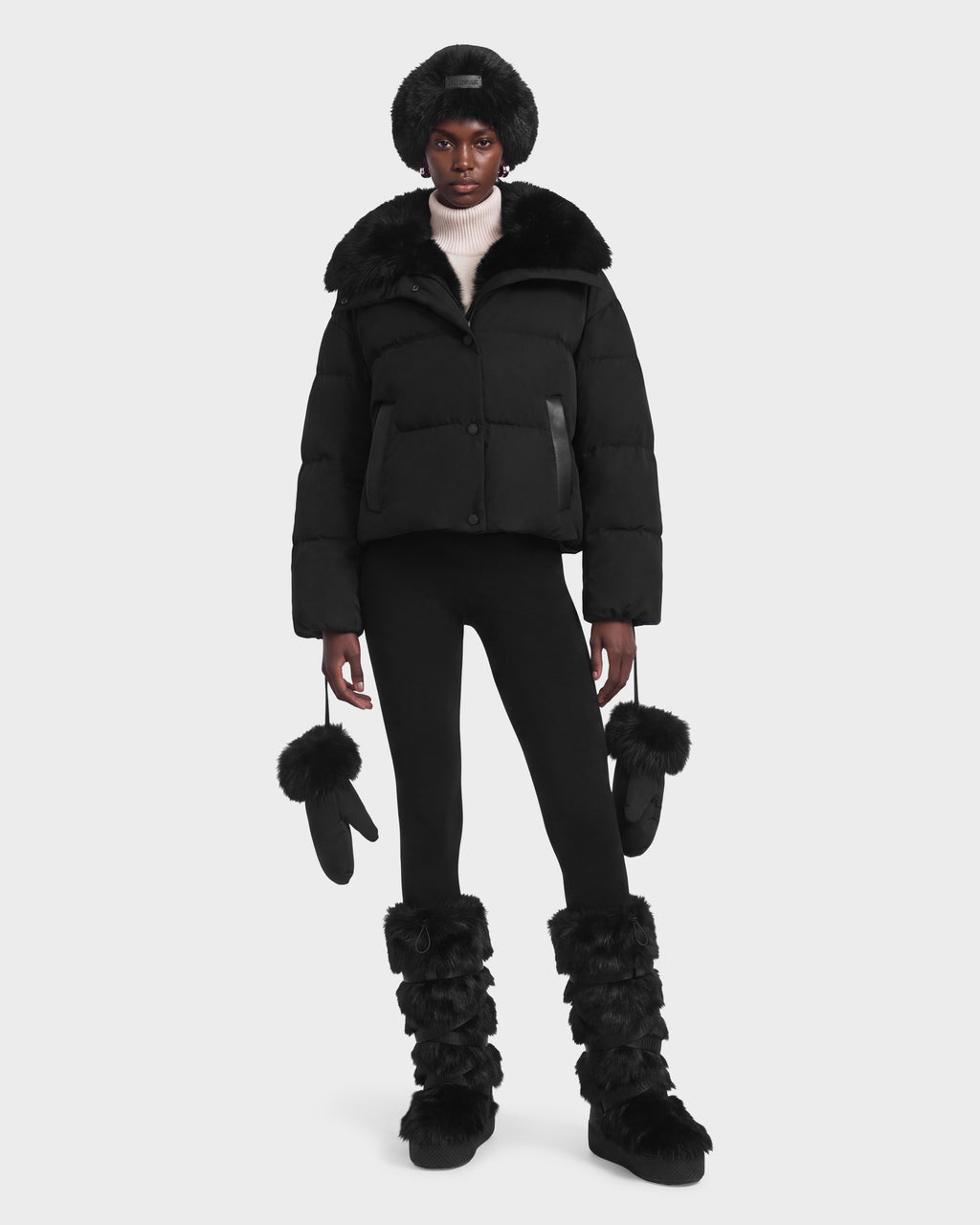 A model wearing a black Rudsak ALONDRA - 8126992 Black puffer jacket with a fur-trimmed collar, black leggings, a fur hat, fur-trimmed mittens, and fuzzy black boots.