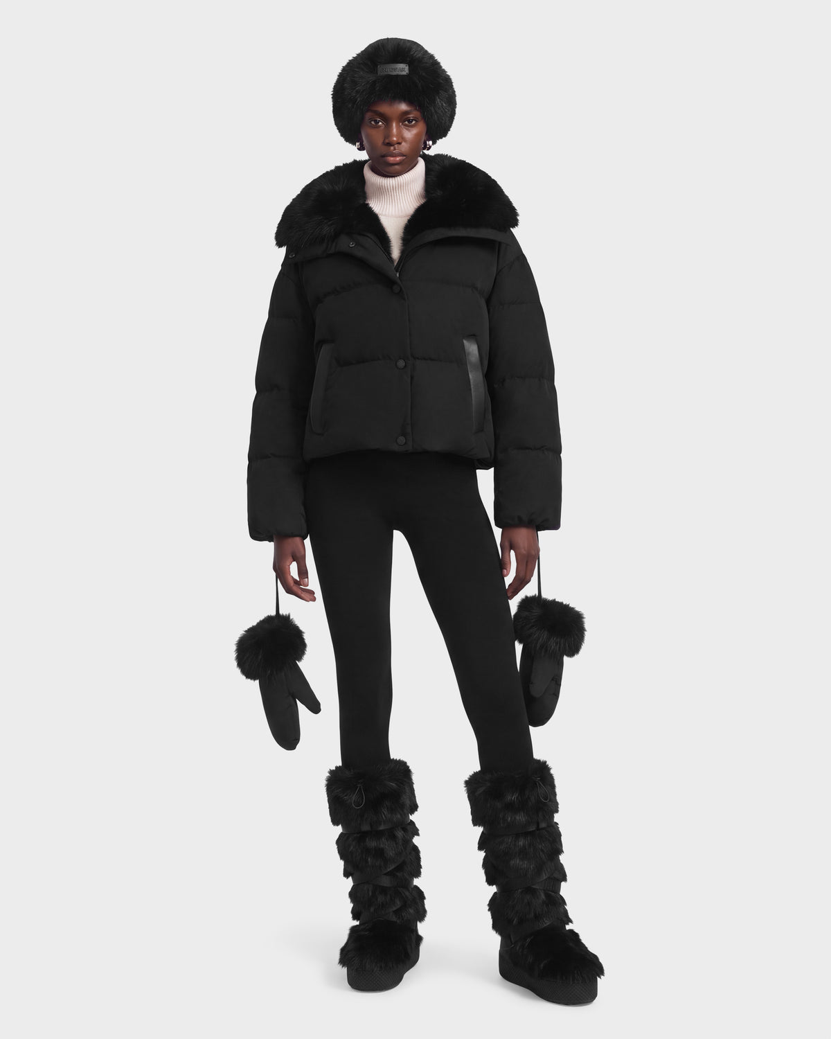 A model wearing a black Rudsak ALONDRA - 8126992 Black puffer jacket with a fur-trimmed collar, black leggings, a fur hat, fur-trimmed mittens, and fuzzy black boots.