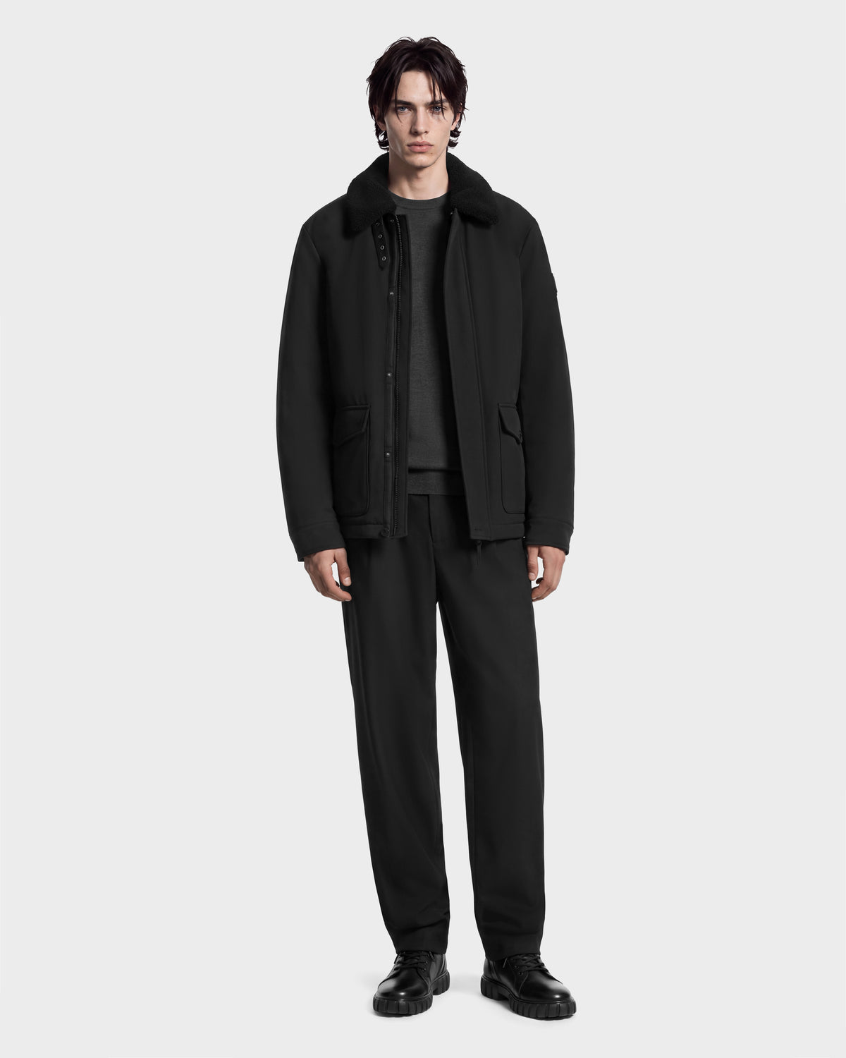 A man wearing the Rudsak ANTOINE - 6126506 Black utility jacket with a black faux-fur collar over a grey sweater, paired with black trousers and black shoes.