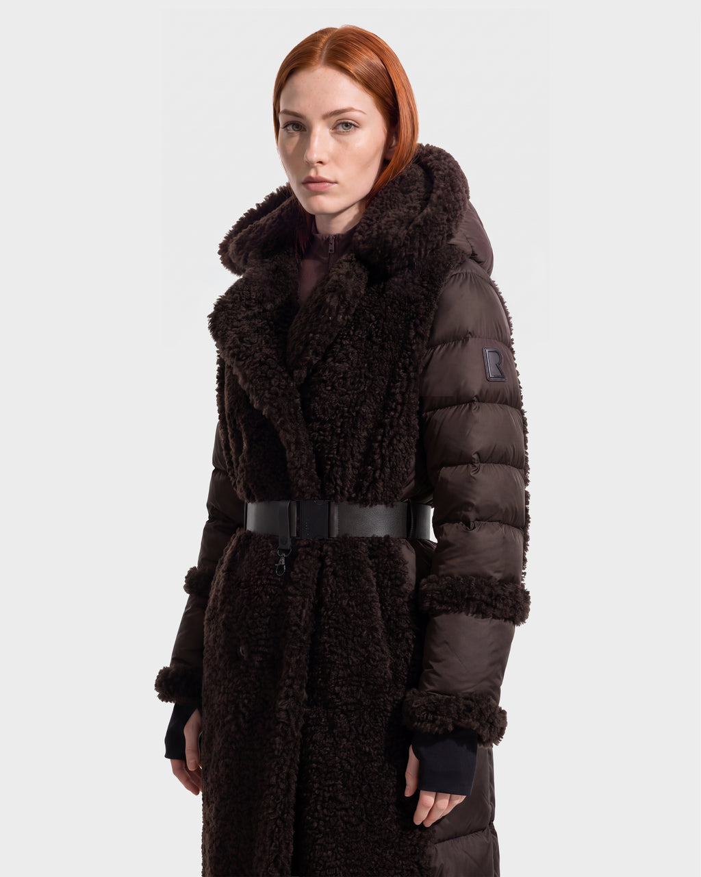 A person with red hair wears the Rudsak NOELLE - 8126971 Espresso, a long, dark brown (espresso) hooded winter coat featuring a thick collar, black belt, and logo patch on the sleeve, standing against a plain white background.