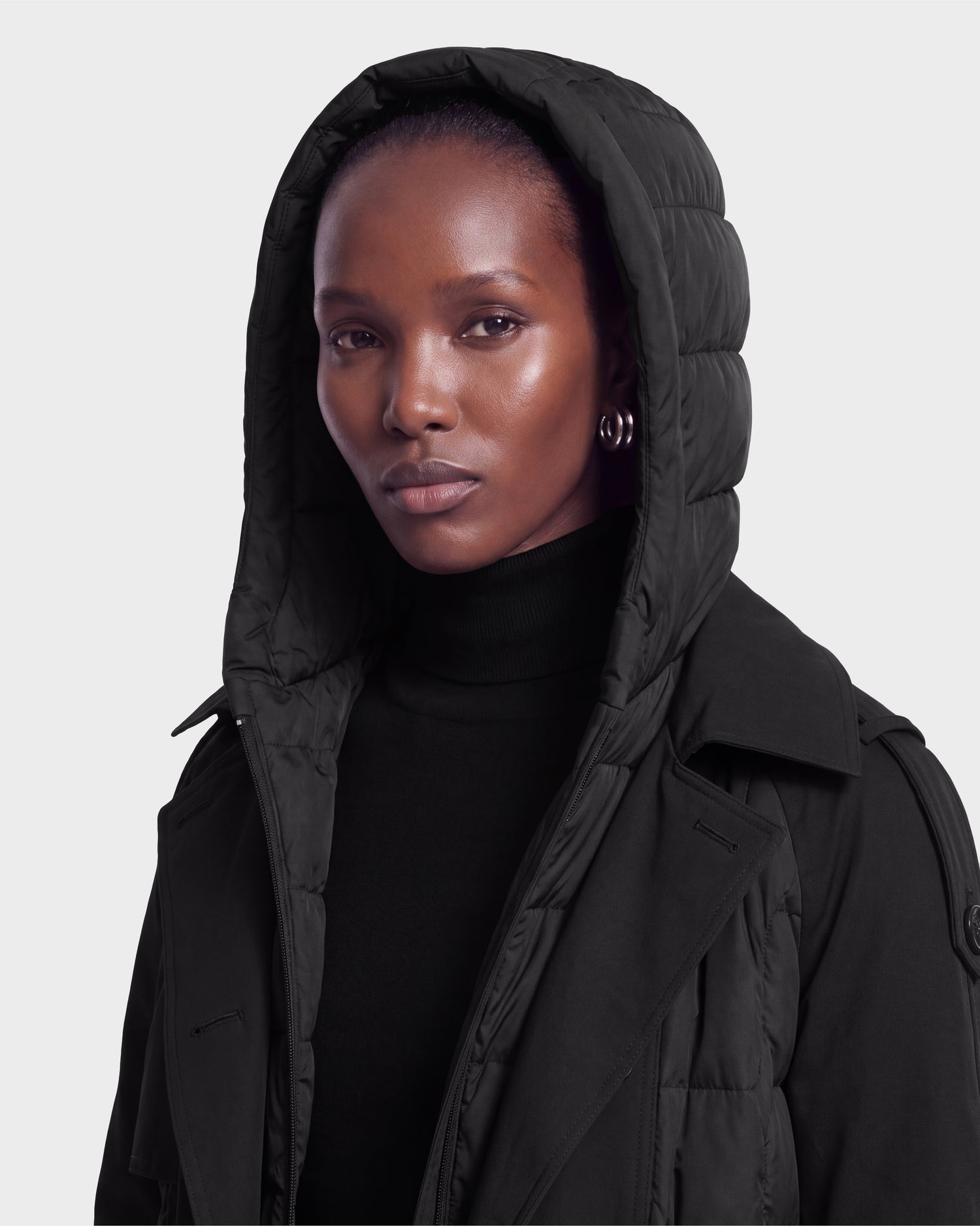 A person wearing the Rudsak ELVIE - 8126512 Black hooded puffer jacket over a black turtleneck looks at the camera with a neutral expression against a plain light background.
