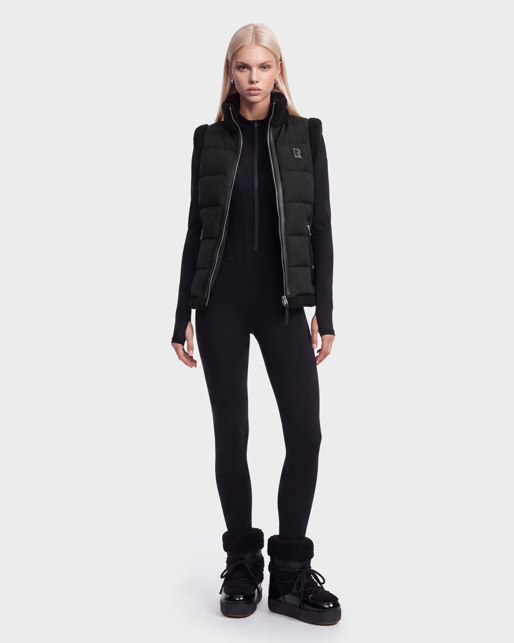 A person with long blonde hair wears the Rudsak HALI - 8126771 Black puffer vest filled with RDS-certified down, styled with a black fitted jumpsuit and black winter boots, standing against a plain white background.