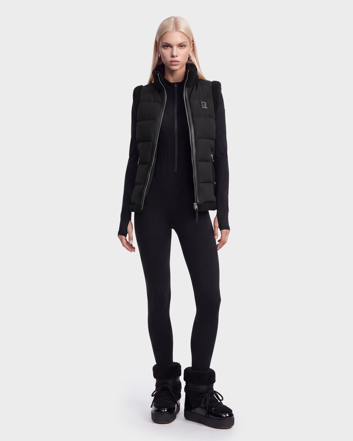A person with long blonde hair wears the Rudsak HALI - 8126771 Black puffer vest filled with RDS-certified down, styled with a black fitted jumpsuit and black winter boots, standing against a plain white background.