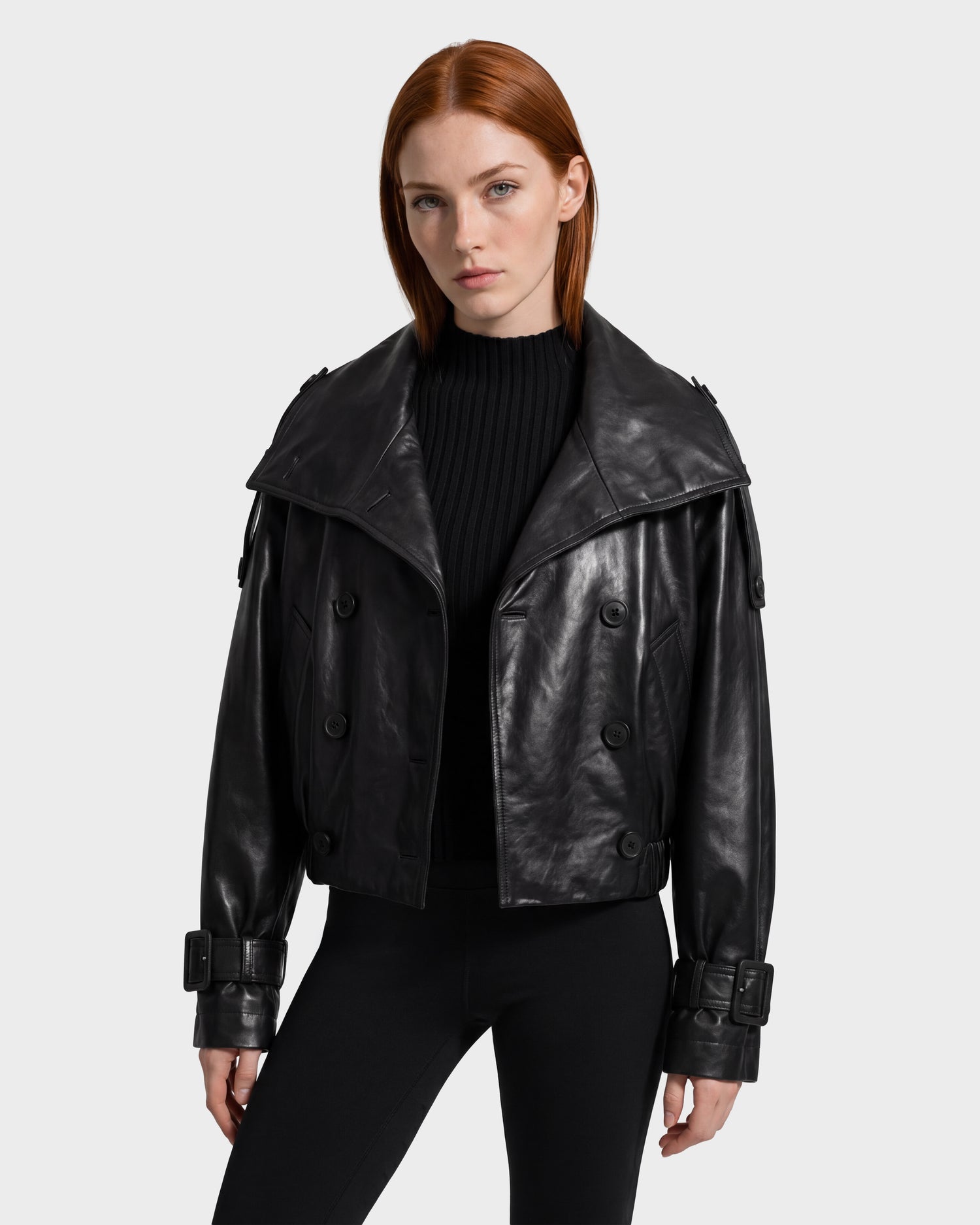 A person with straight red hair wears the Rudsak JANELLE - 8126936 Black leather bomber jacket, featuring wide lapels, buckle cuffs, and a modern silhouette, layered over a black ribbed sweater and black pants while standing against a plain light background.