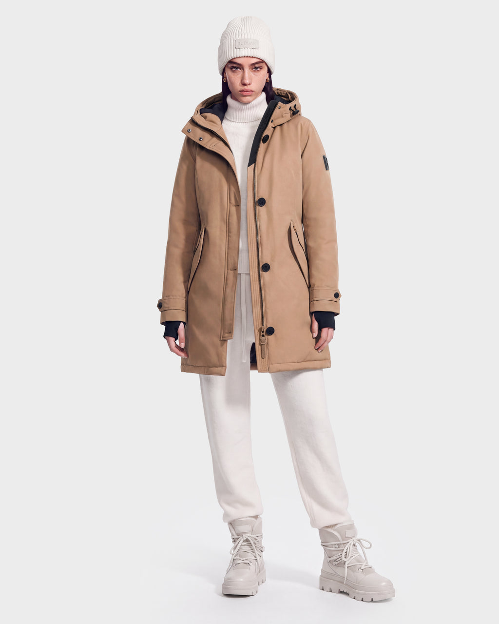A person wearing the Rudsak KEYSHIA - 8126517 Camel parka, white turtleneck, white knit beanie, white joggers, and beige boots stands against a plain white background.