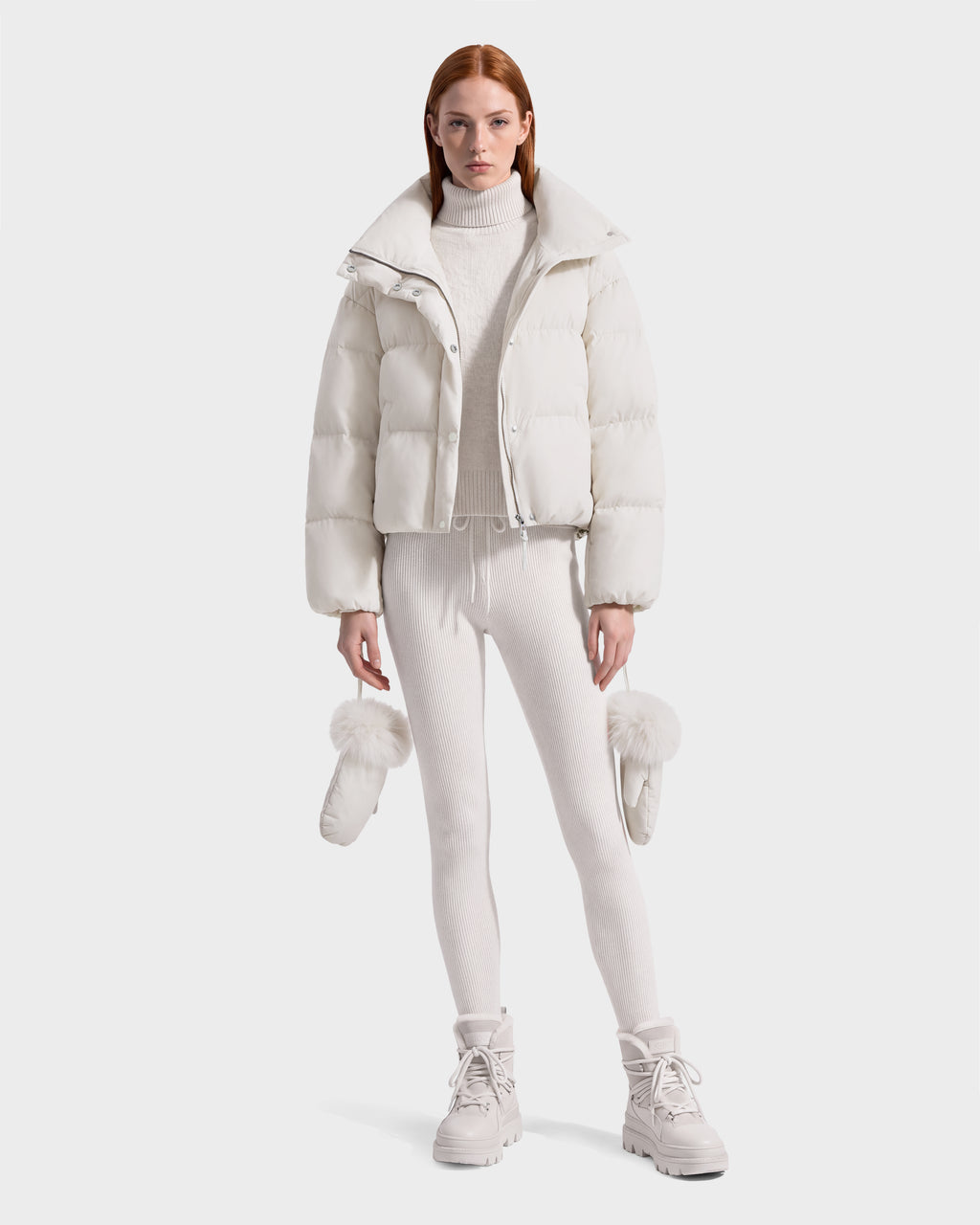 Model wearing an ALONDRA - 8126992 Vanilla white cropped puffer jacket, matching turtleneck sweater and leggings, holding a pair of fur-trimmed mittens.