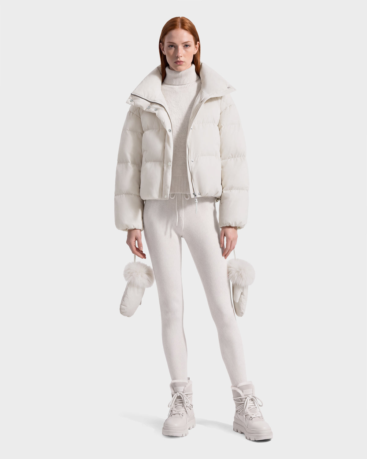 Model wearing an ALONDRA - 8126992 Vanilla white cropped puffer jacket, matching turtleneck sweater and leggings, holding a pair of fur-trimmed mittens.