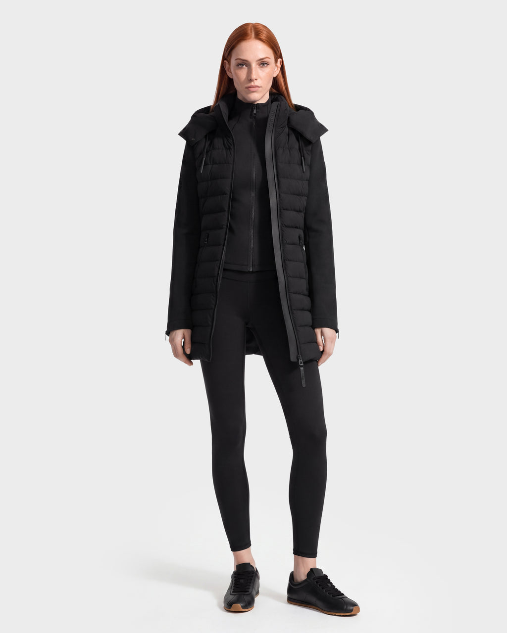 A woman with long red hair wears the Rudsak MERINA - 8125056 Black women’s light jacket—a black padded coat—paired with black leggings, a black zip-up top, and black sneakers, standing against a plain white background.