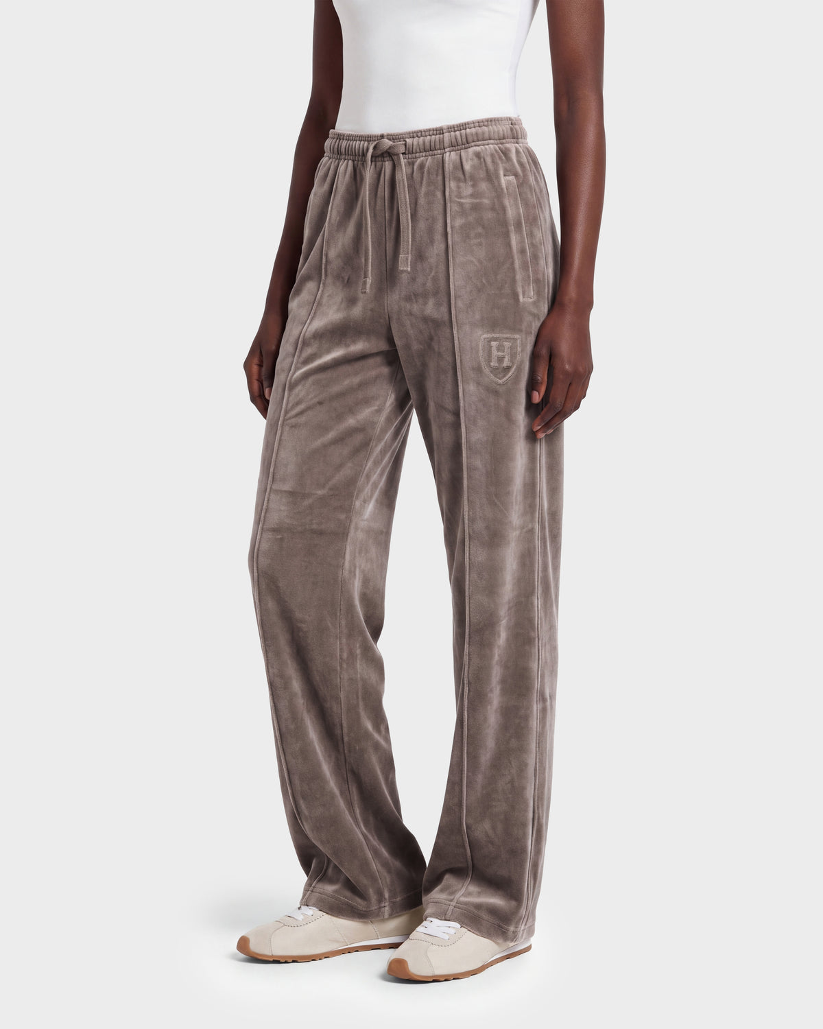 A person stands against a plain background wearing Rudsak’s OLIVYA HV - H526903 Taupe velour drawstring pants with a crest on the hip, paired with a white tank top and beige sneakers.