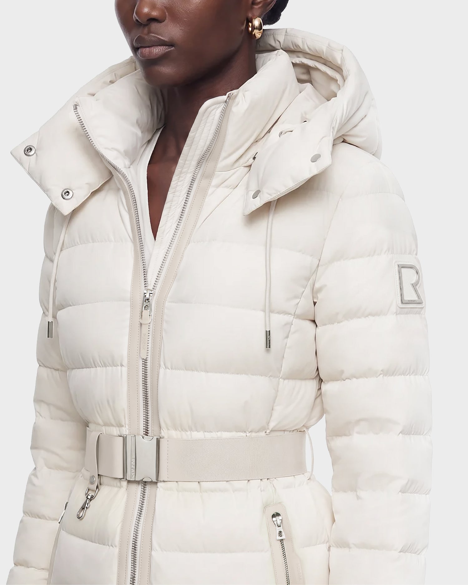A woman wearing the Rudsak PELIN S - 8124813 Vanilla quilted jacket, featuring a belt, hood, high collar, and an upper sleeve patch.