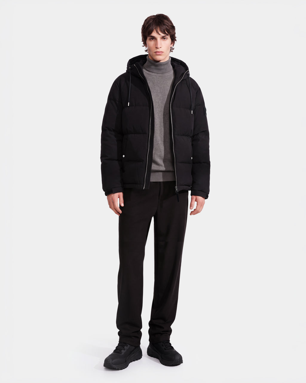 A person wearing the Rudsak STERLING EL - 6126759 Black puffer jacket over a gray turtleneck sweater and black pants stands against a plain white background.