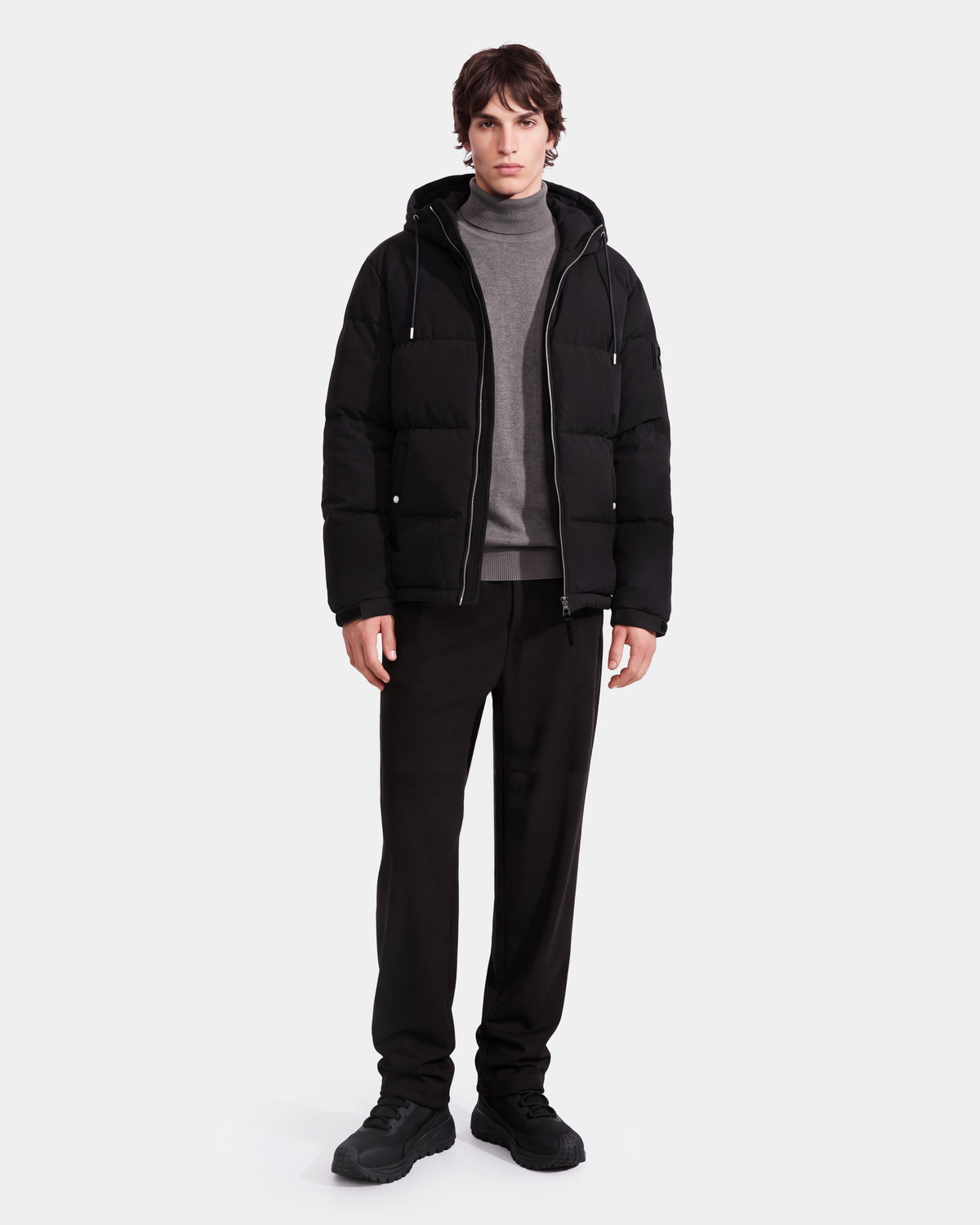 A person wearing the Rudsak STERLING EL - 6126759 Black puffer jacket over a gray turtleneck sweater and black pants stands against a plain white background.