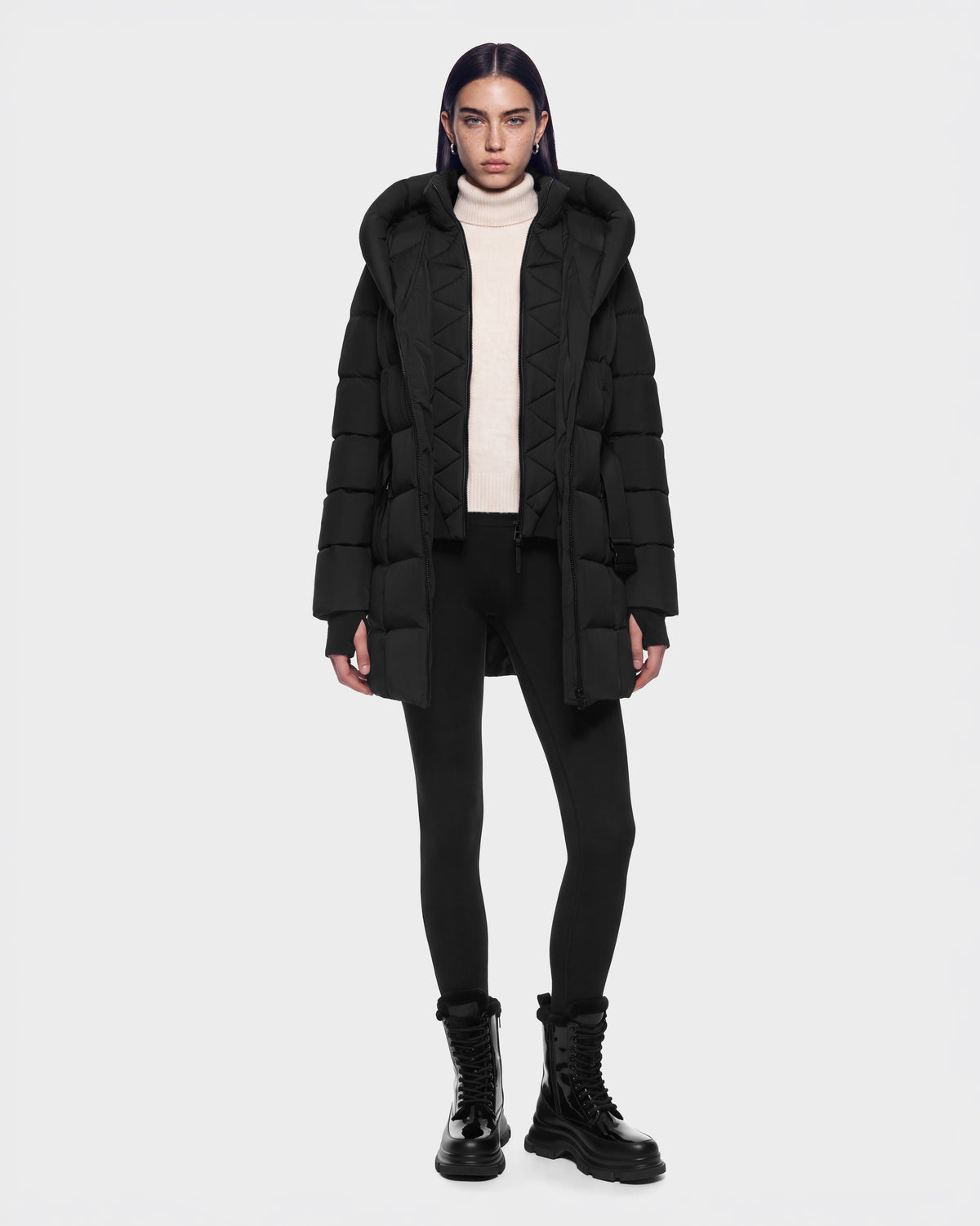 A person wearing a RUDSAK TAYLOR M - 8125965 Black hooded puffer jacket over a cream turtleneck, black leggings, and black lace-up boots.