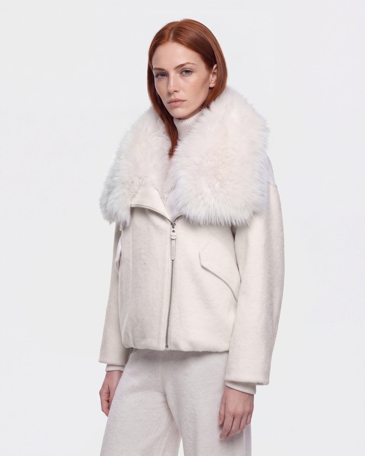 A woman with long red hair wears the Rudsak ZARIA - 8126999 Vanilla white winter puffer jacket, featuring a large, fluffy faux-fur collar, paired with white pants as she stands against a plain white background.