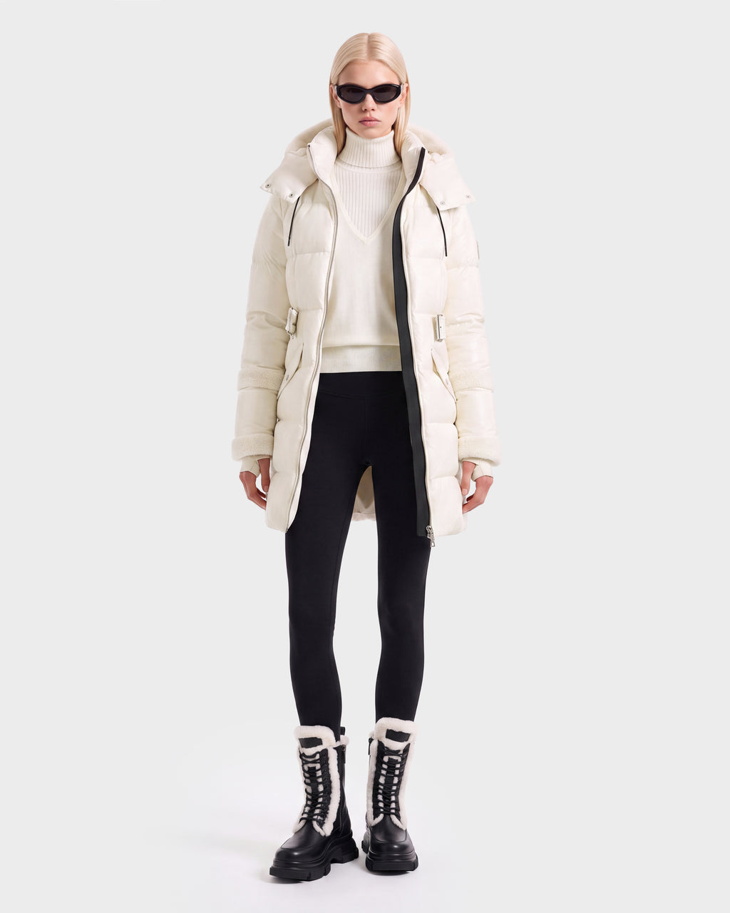 A model wears a knee-length vanilla AUDREY L - 8124585 puffer jacket made from recycled Stormshell fabric over a white sweater, black leggings, and black fur-lined lace-up winter boots.