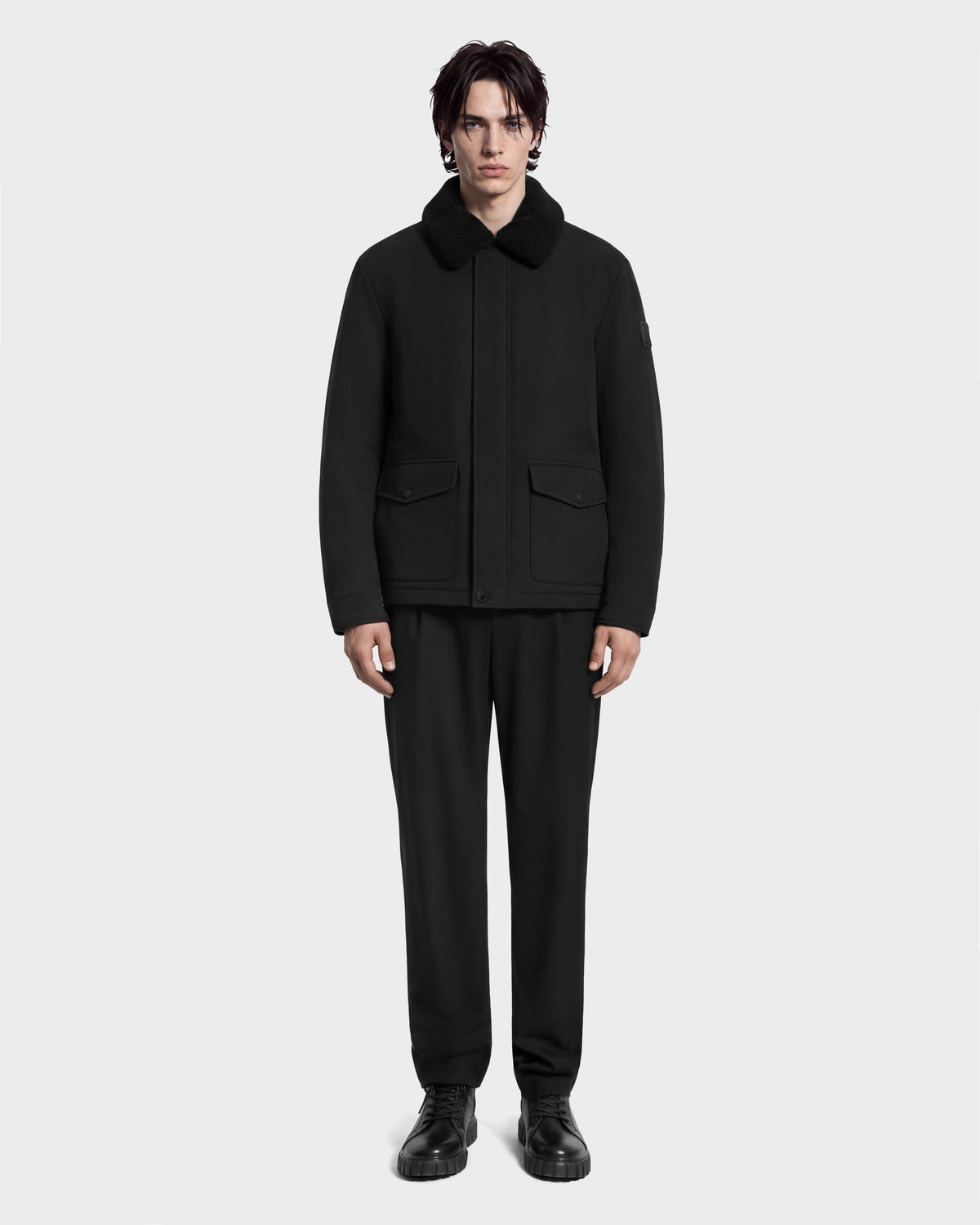 A man wearing an ANTOINE - 6126506 Black refined utility jacket with a faux-fur collar, paired with matching black trousers and boots.
