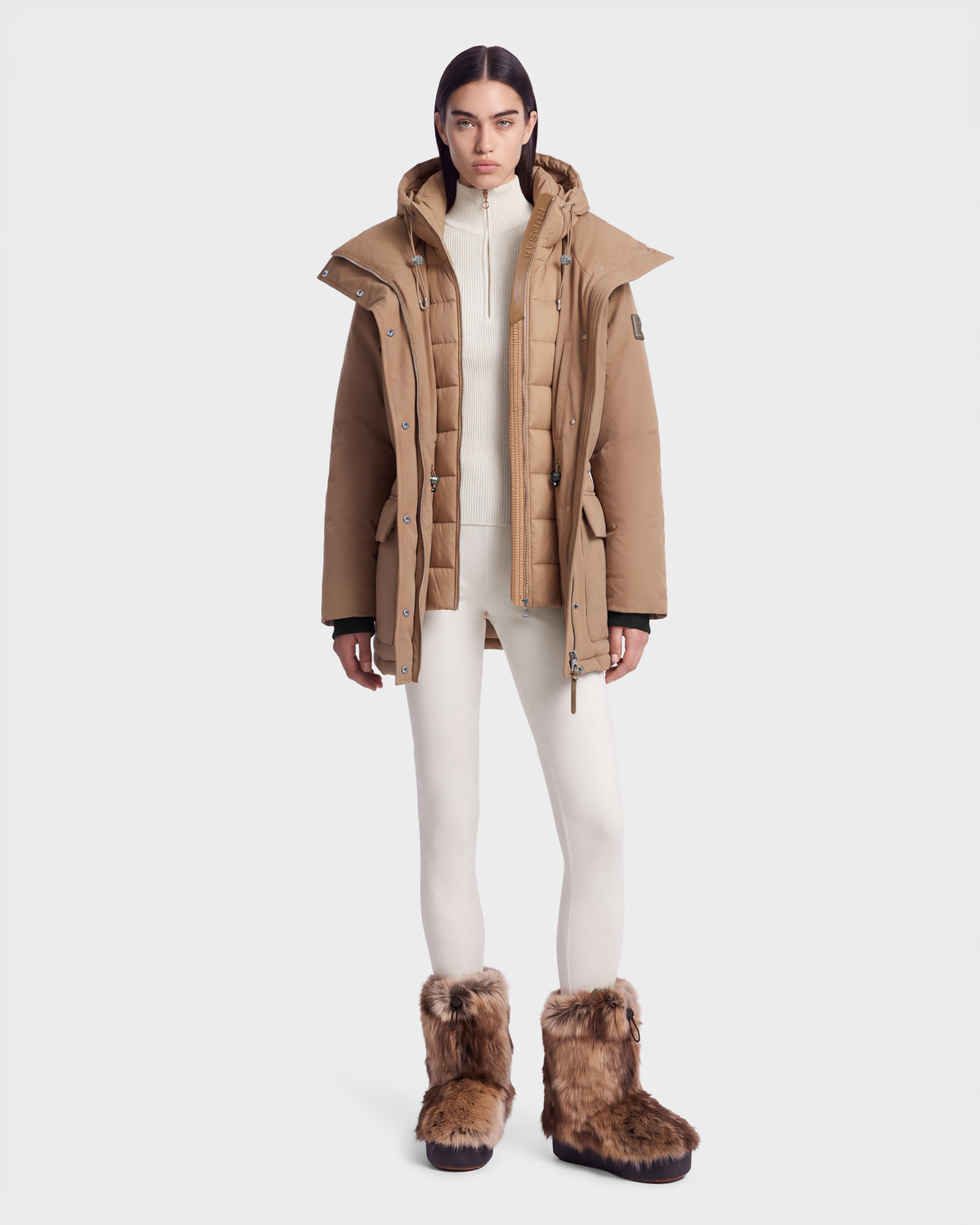 A person wears a camel-colored Rudsak BHIMA - 8126515 Camel hooded down parka over a white quarter-zip sweater and white leggings, paired with brown faux-fur trimmed winter boots.