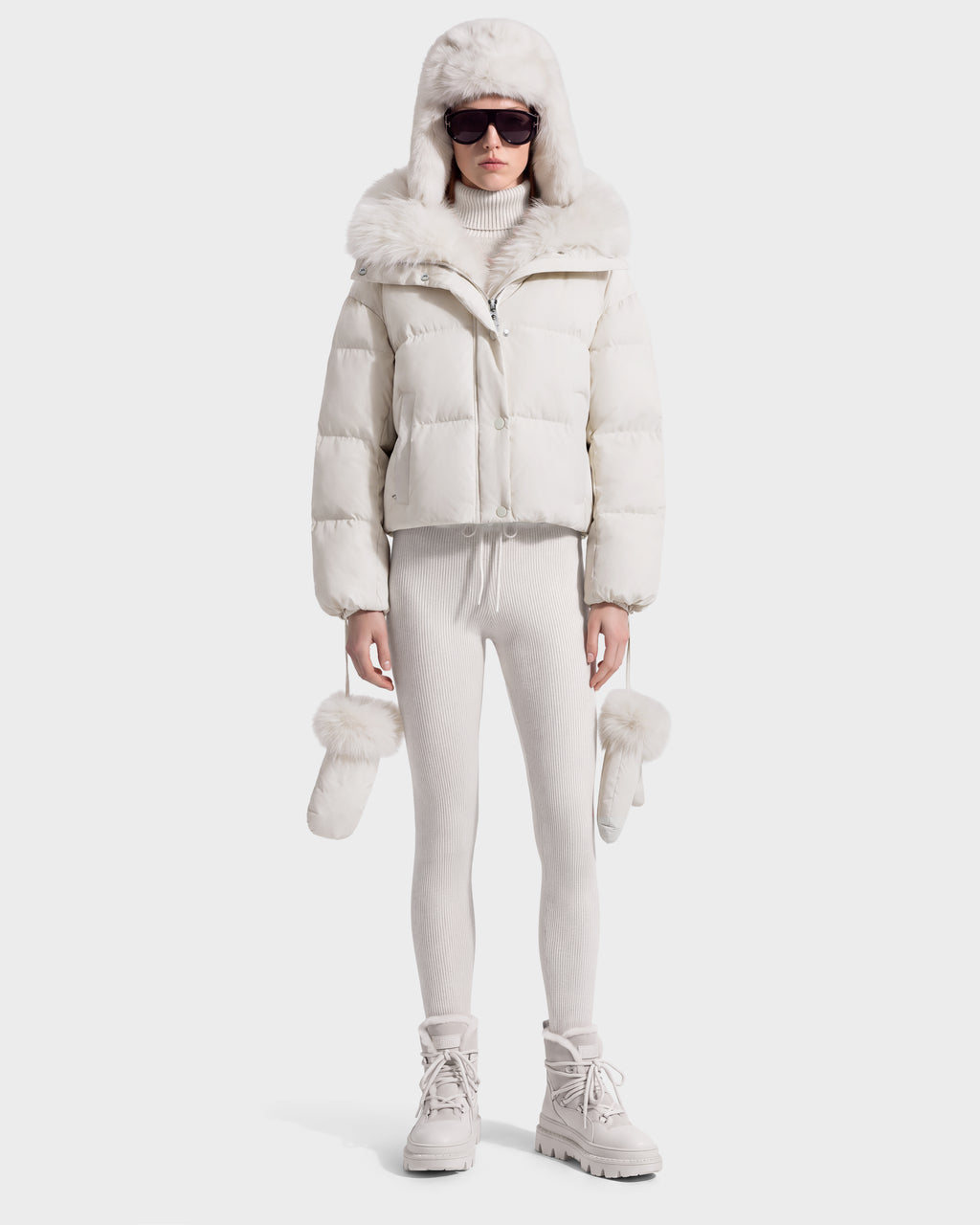 A person wearing a Rudsak ALONDRA - 8126992 Vanilla cropped white puffer jacket, fur-trimmed trapper hat, sunglasses, matching knit leggings, and winter boots, holding a pair of fur-accented mittens.