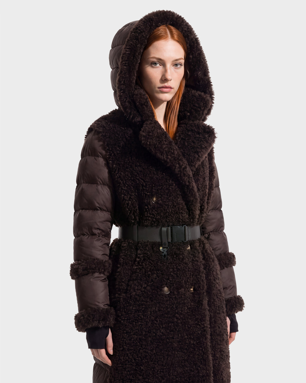 A woman with straight red hair wears the Rudsak NOELLE - 8126971 Espresso, a dark brown hooded winter coat with a textured faux fur front and sleeves, belted waist, and snap buttons. She faces forward against a plain, light background.