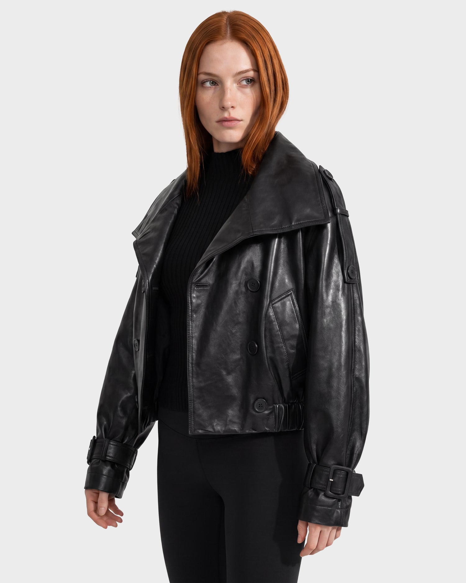 A woman with straight red hair wears the Rudsak JANELLE - 8126936 Black leather bomber jacket, featuring large lapels, buckled cuffs, and a modern silhouette. She pairs it with a black top and pants while standing against a plain light gray background.