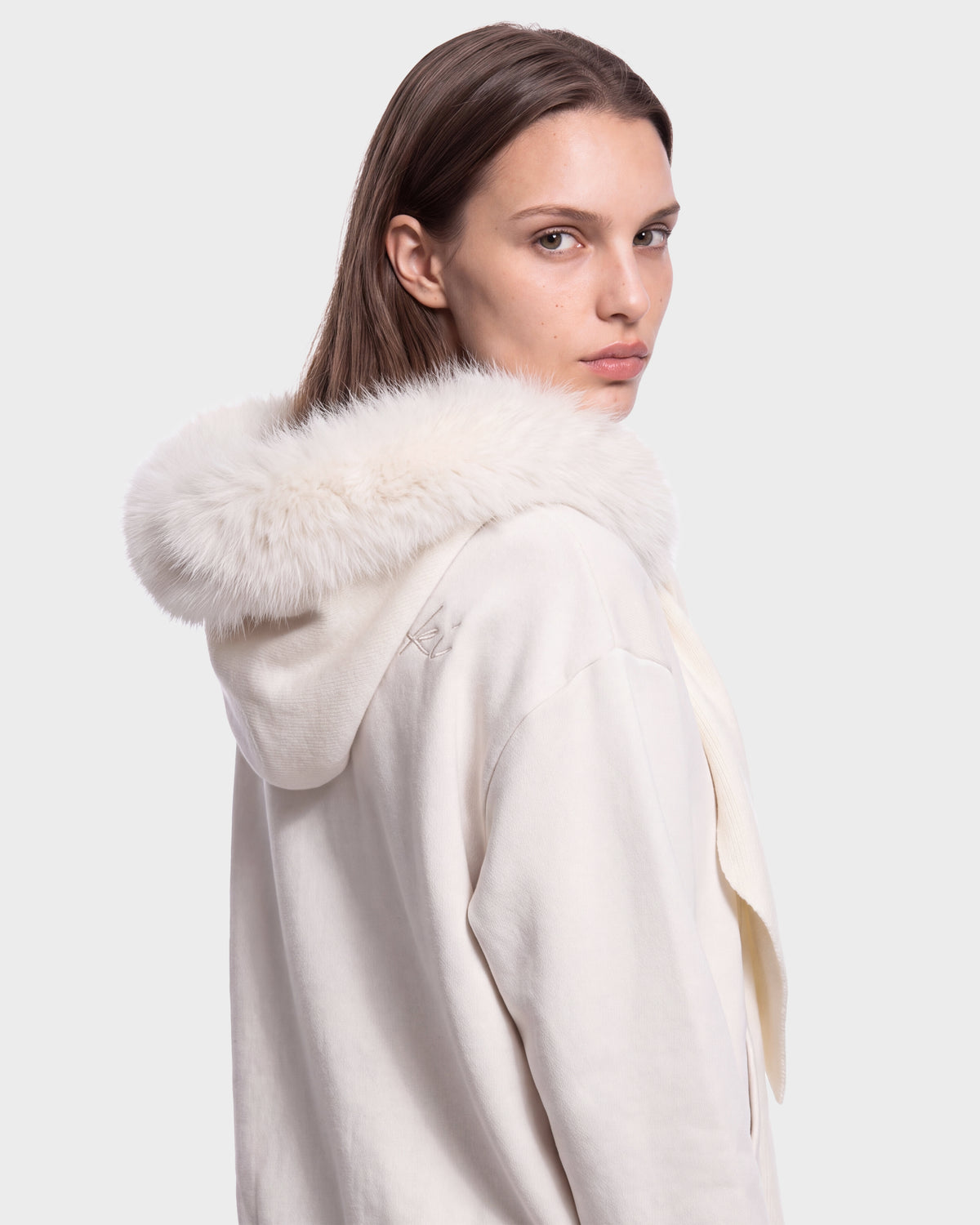 A person with straight brown hair looks over their shoulder, wearing the Rudsak KARYMA - 8426507 Vanilla cream-colored hoodie featuring a fluffy white fur trim on the hood. The background is plain and light gray.