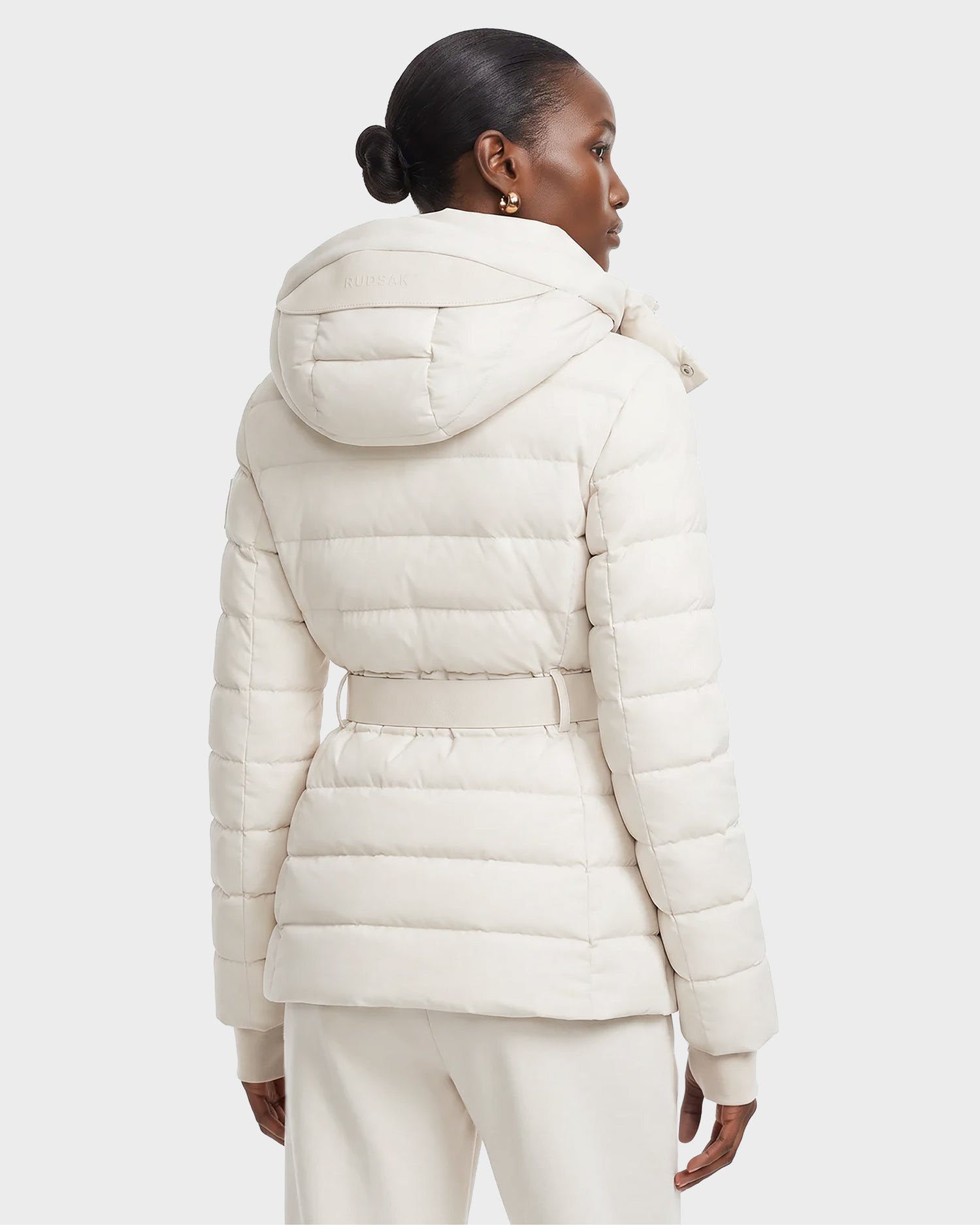 A woman with her hair in a low bun wears the Rudsak PELIN S - 8124813 quilted jacket in Vanilla, featuring a belt and hood, as she faces away from the camera against a plain background.