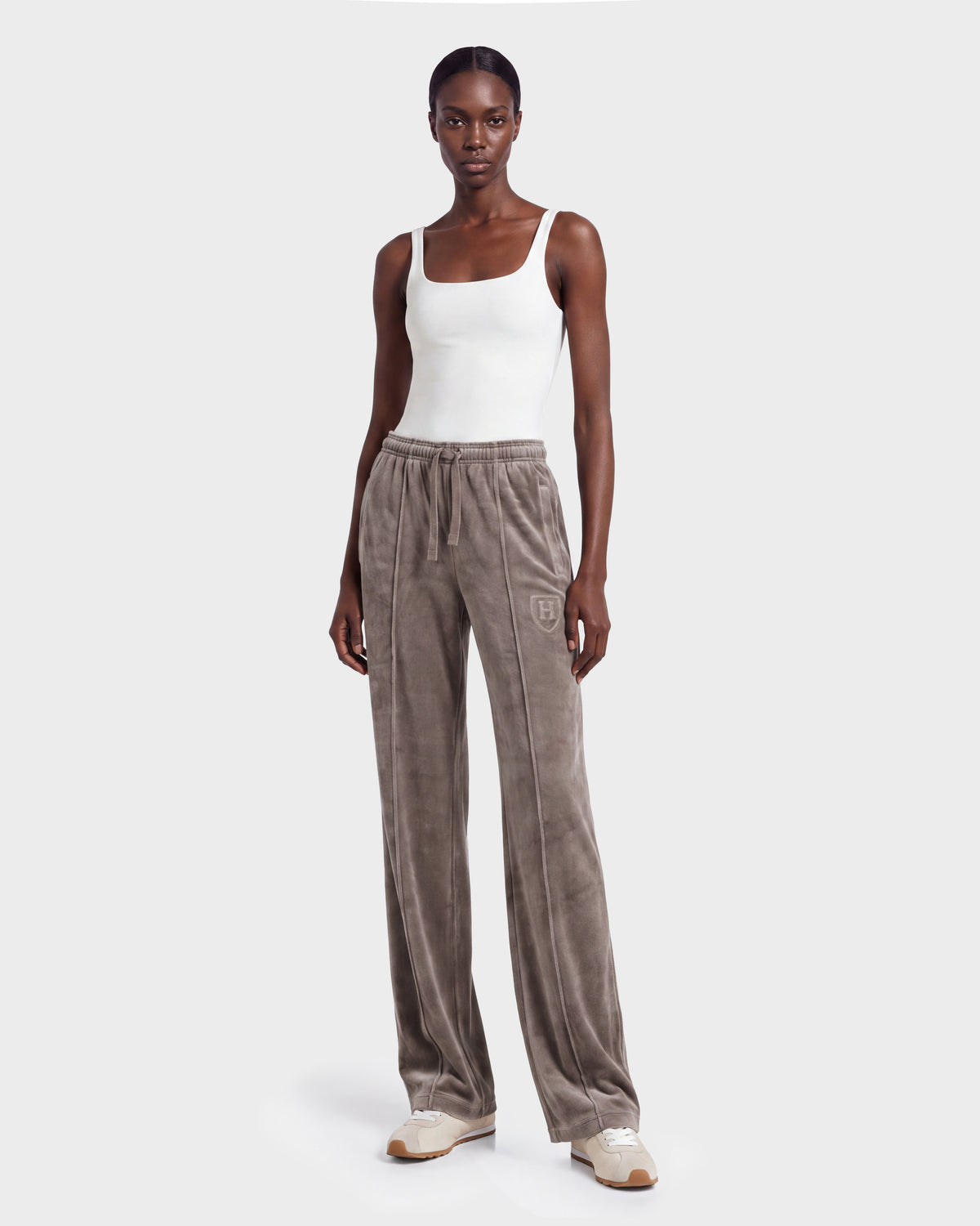 A woman wearing a white tank top, Rudsak OLIVYA HV - H526903 Taupe velour wide-leg sweatpants with a logo, and white sneakers stands against a plain light gray background.
