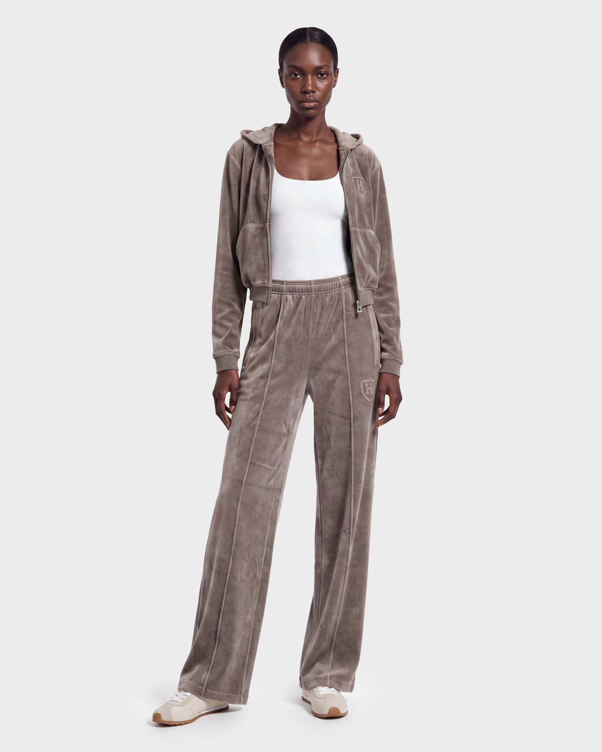 A woman stands against a plain light background wearing the Rudsak TYRA HV - H526904 Taupe velour tracksuit, featuring a zip-up hoodie and matching wide-leg pants, styled with a white tank top and beige and white sneakers.