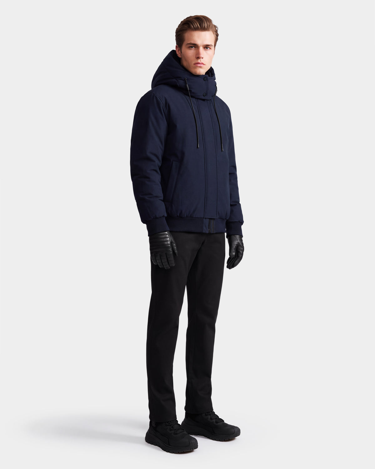 A man stands against a plain white background wearing the Rudsak WILL - 6126561 Navy RDS-certified down bomber jacket, paired with black pants, black gloves, and black shoes.