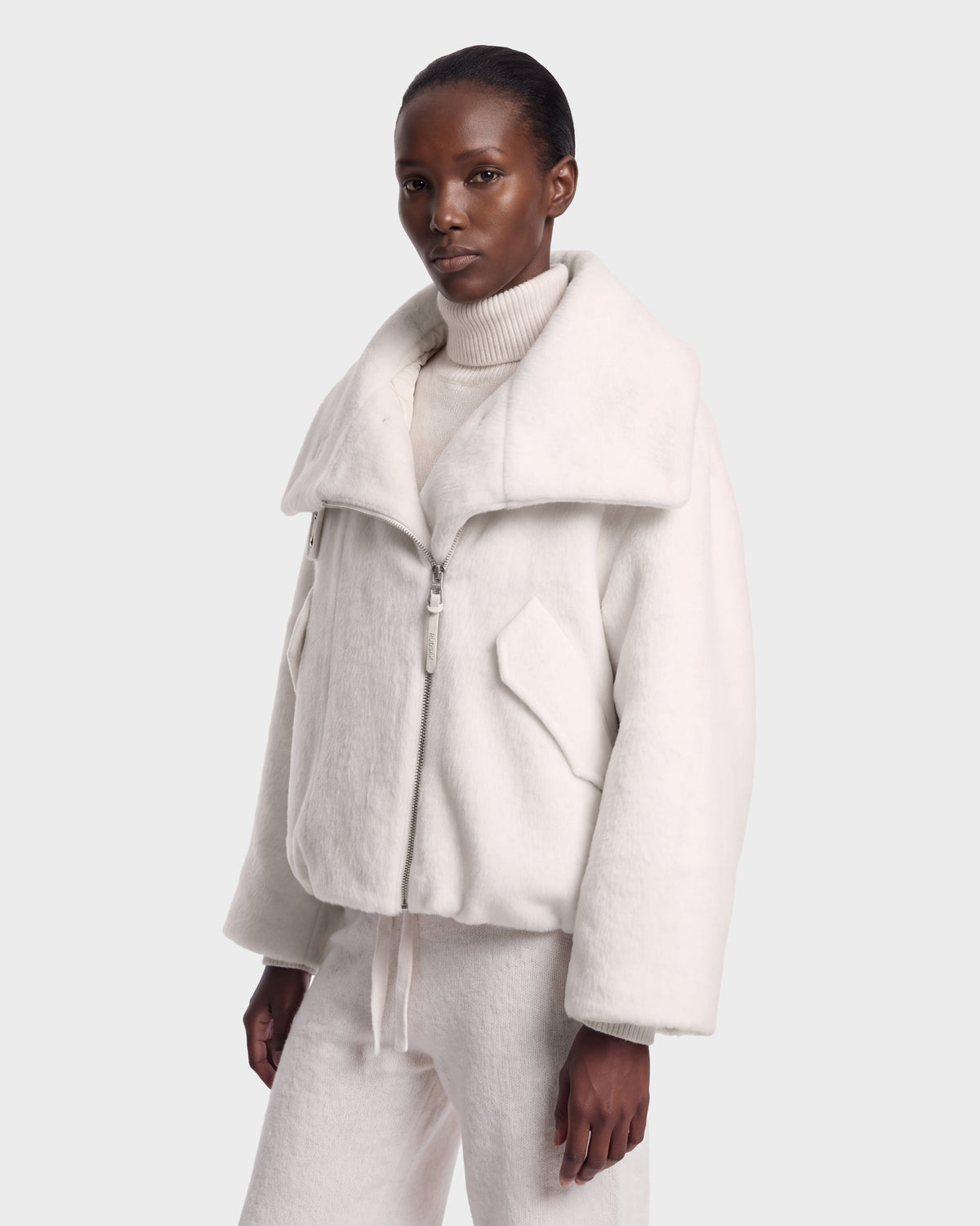 Wearing the Rudsak ZARIA - 8126999 Vanilla, an oversized white zip-up jacket with a wide collar, layered over a white turtleneck and matching white pants, the person stands against a plain light gray background.
