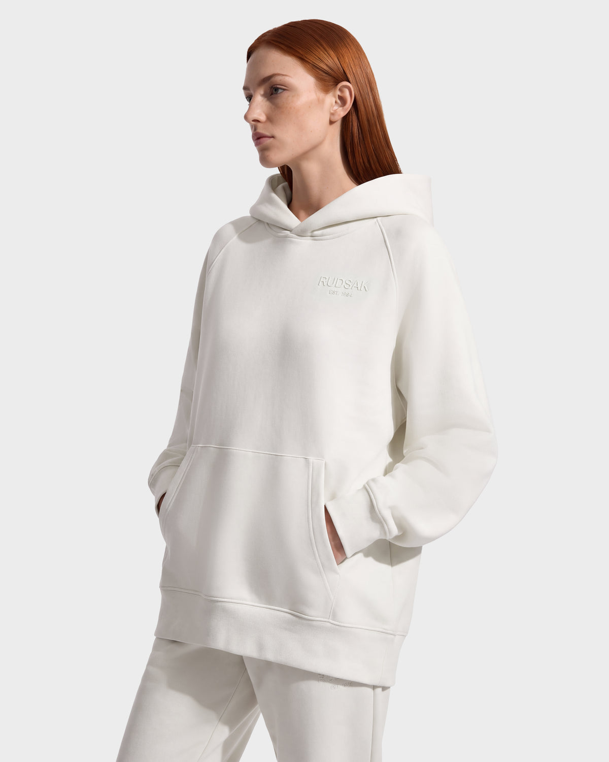 A person wearing the Rudsak BRIAR - 9525001 Vanilla W, an oversized cream-colored unisex hoodie with a kangaroo pocket and subtle embossed branding on the chest, perfect for a casual look.
