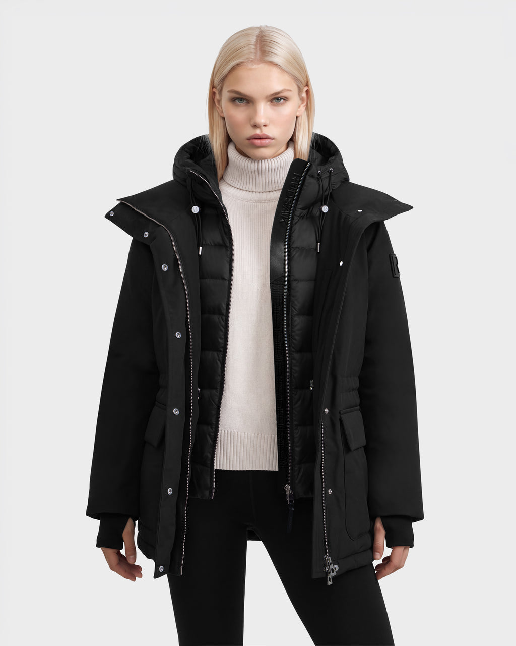 A model wears the BHIMA - 8126515 Black, a hooded parka with a layered front, featuring a quilted inner panel and an outer coat with snap buttons and pockets.