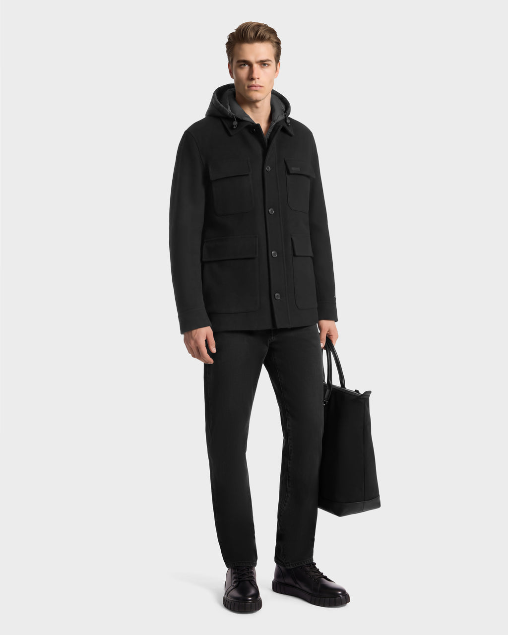 A man wearing a BRUNO - 6126716 Charcoal hooded jacket, black jeans, and black boots, carrying a black tote bag.