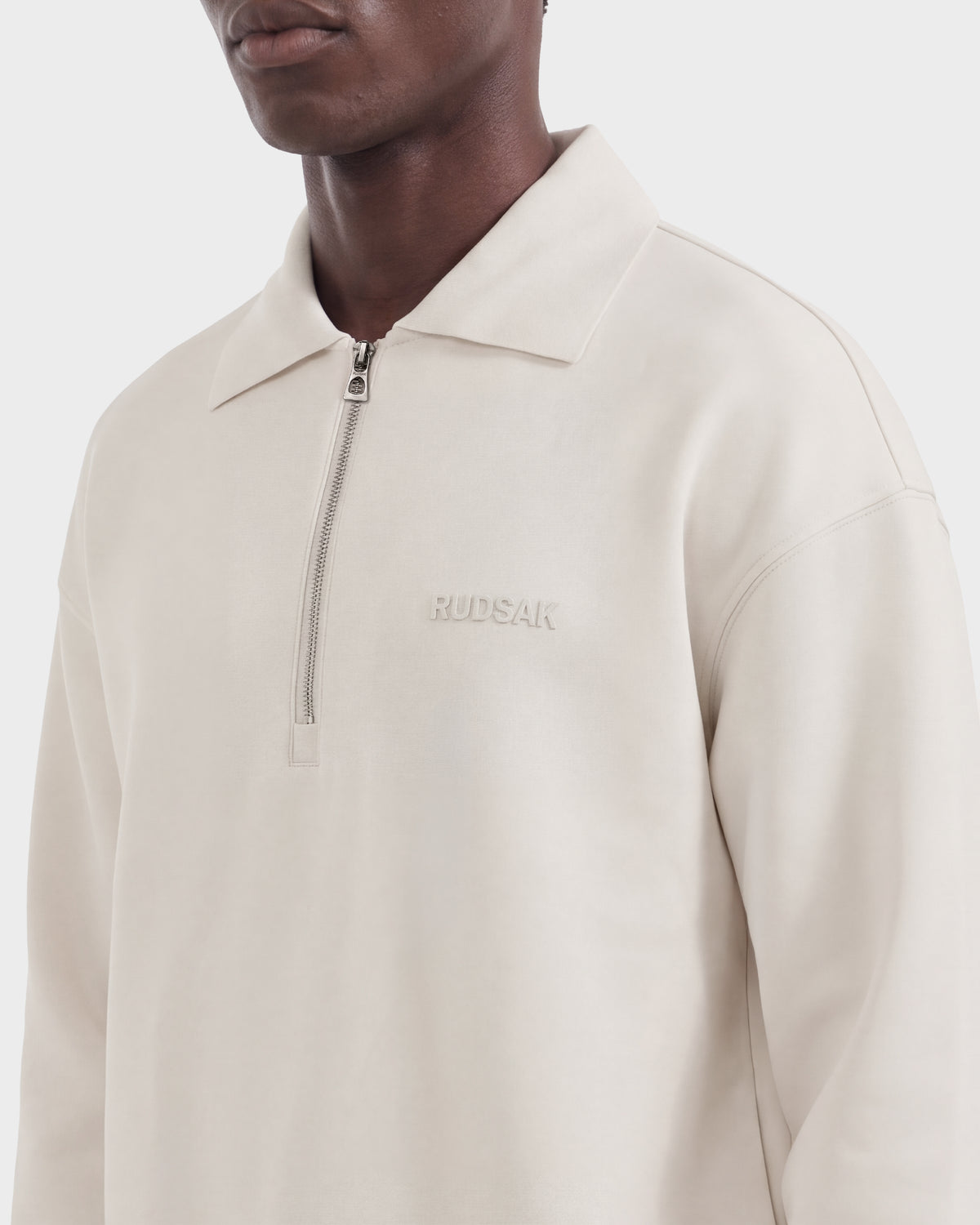 A person wearing the IDRIS - 6526500 Vanilla by Rudsak, a light beige quarter-zip pullover with a collar and RUDSAK embroidered on the left chest. Only the lower face and upper torso are visible.