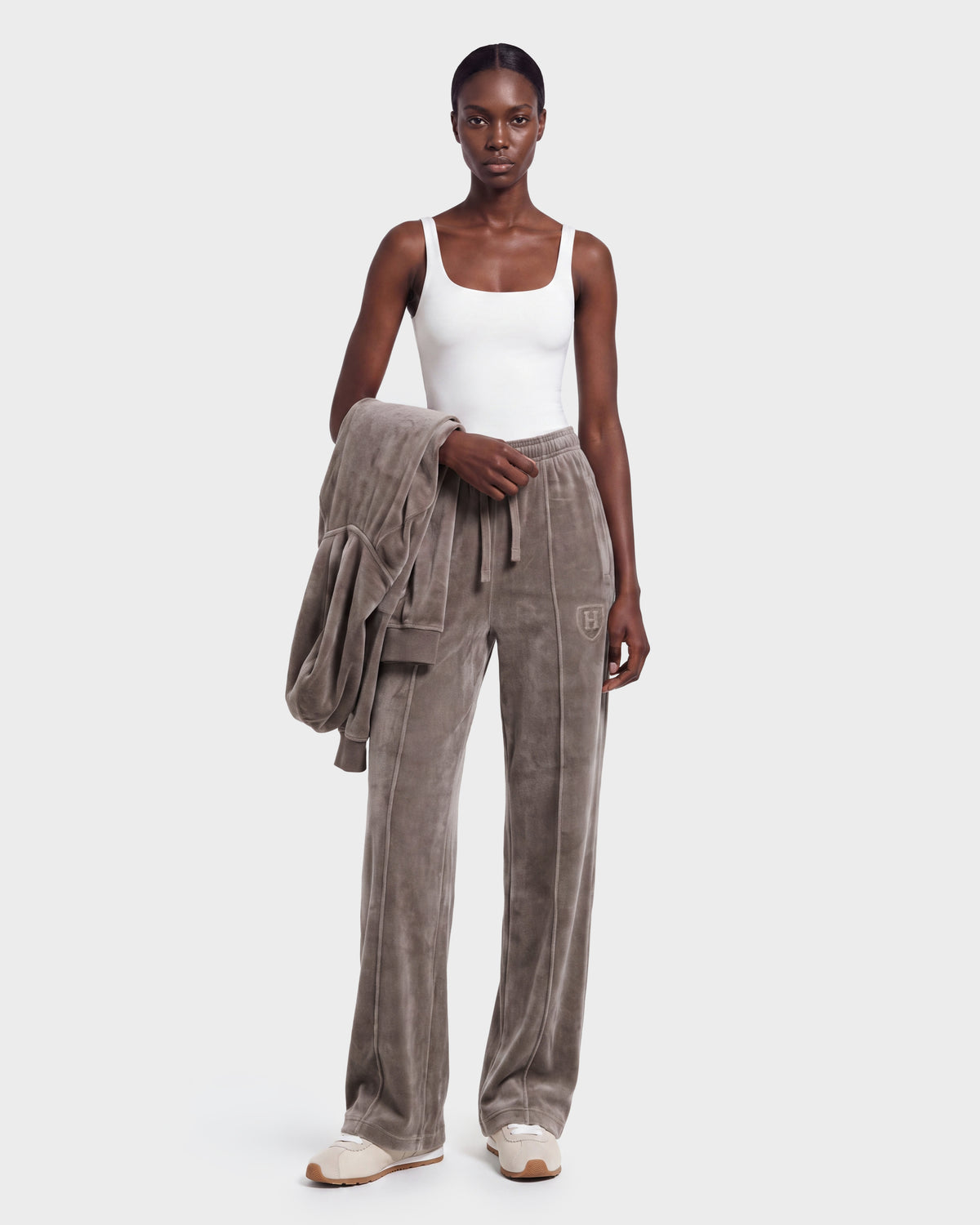 A woman wears the Rudsak OLIVYA HV - H526903 Taupe set, featuring a white tank top and loose gray velour pants, while holding a matching jacket. She completes the look with white sneakers and poses against a plain, light background.