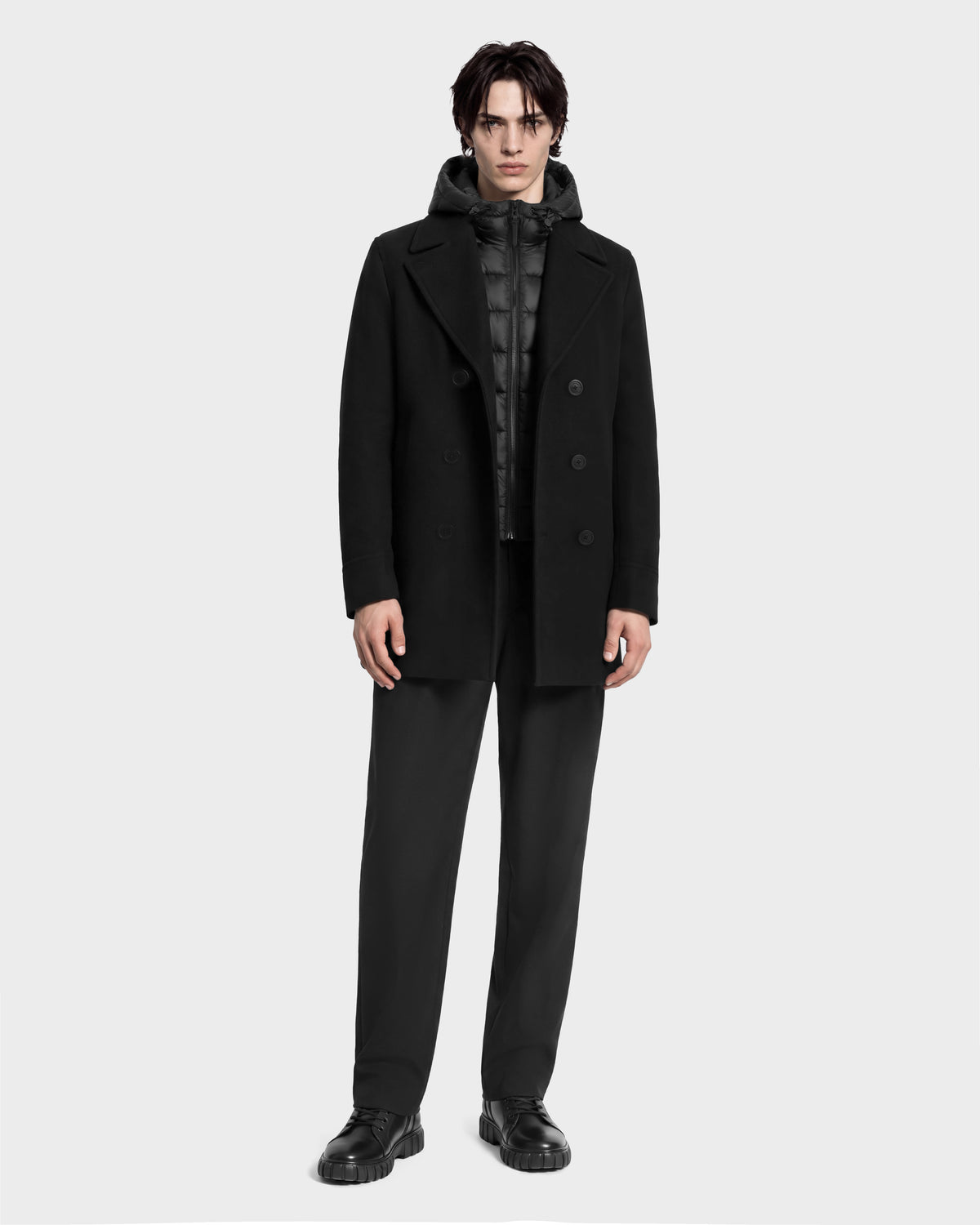 A man wearing a black Rudsak LUCIEN - 6126508 Black wool-blend double-breasted coat over a quilted hooded jacket with duck down insulation, paired with black trousers and black lug-sole shoes.