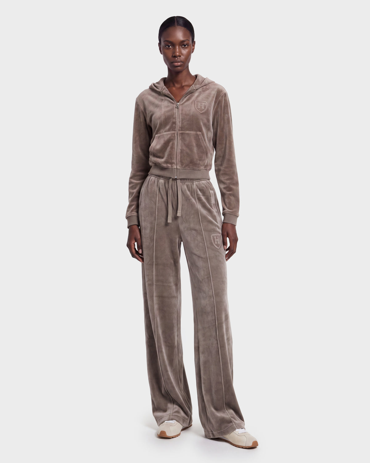 A woman stands against a plain white background, wearing the Rudsak TYRA HV - H526904 Taupe velour tracksuit featuring a zip-up hoodie and wide-leg pants, paired with cream sneakers.