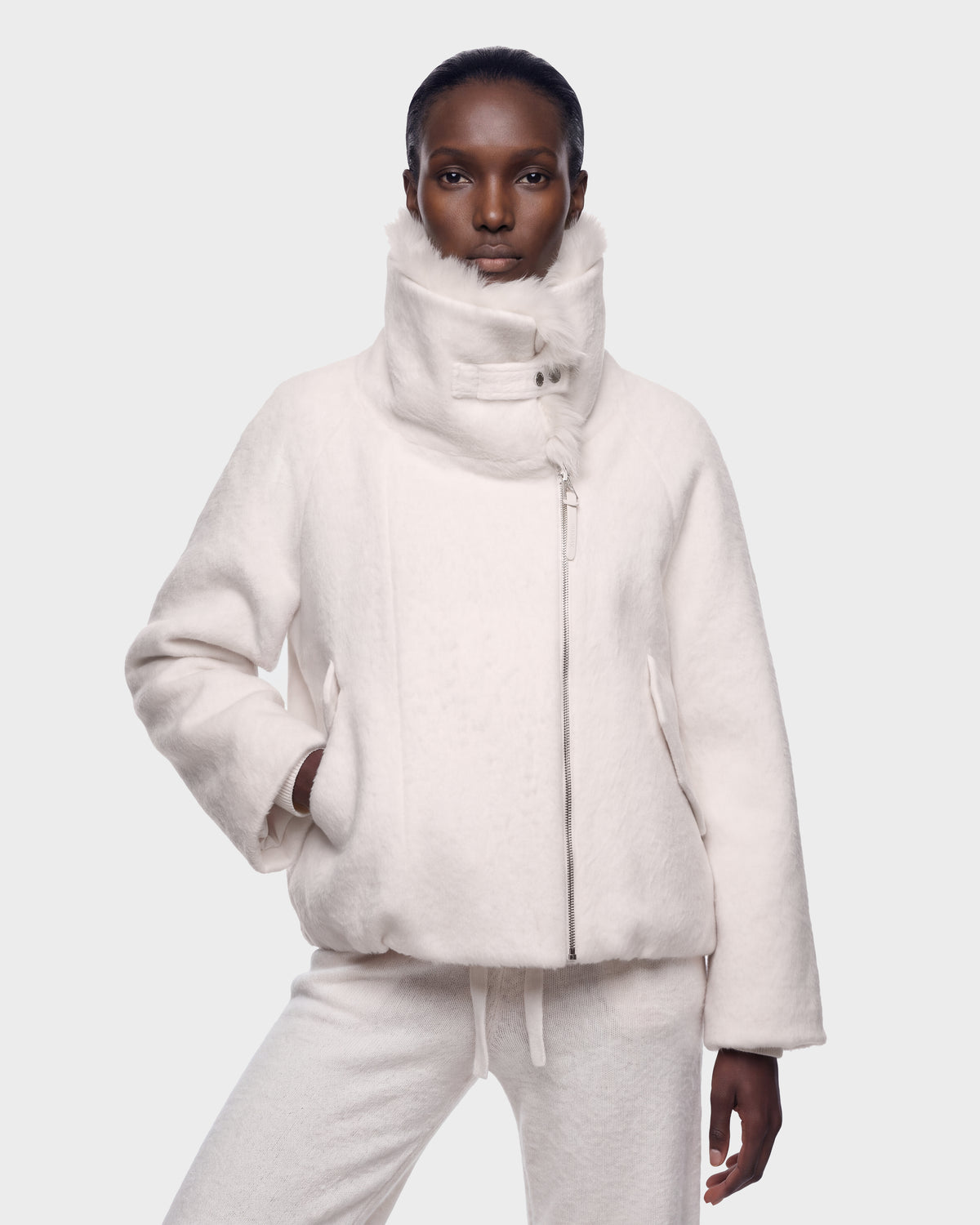 A person stands confidently wearing the Rudsak ZARIA - 8126999 Vanilla high-neck white jacket and matching white pants against a plain light gray background.
