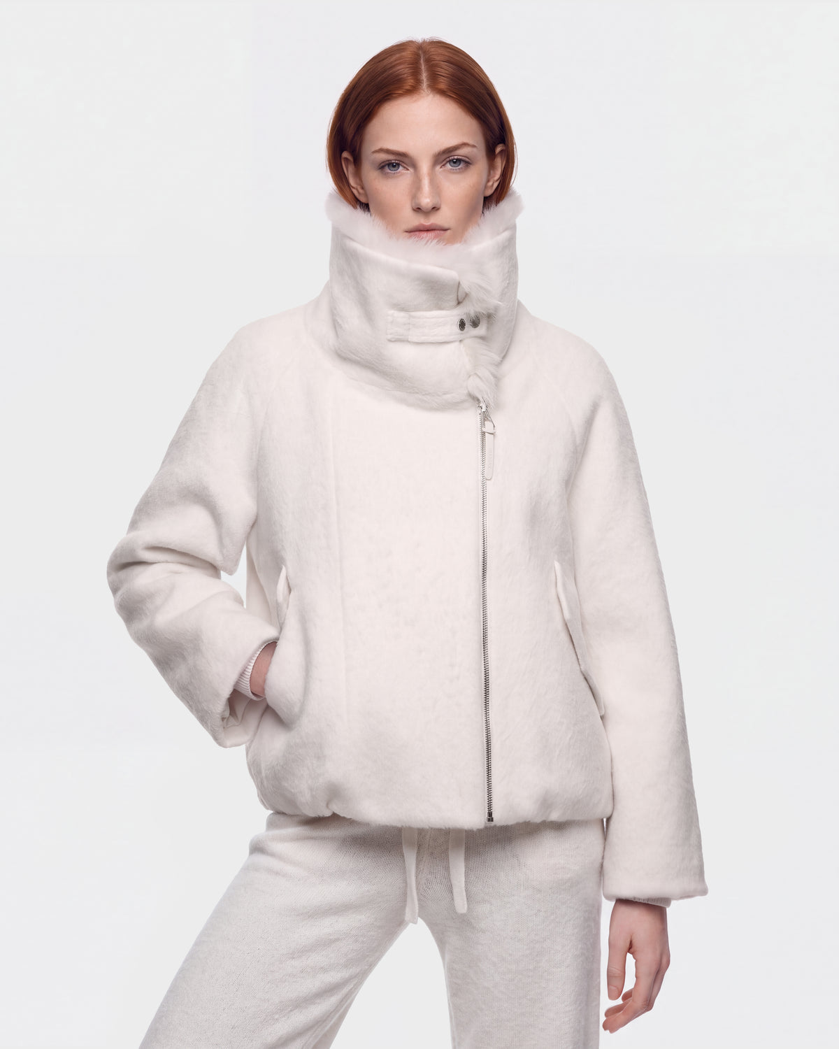 A woman with red hair stands against a plain white background, wearing the Rudsak ZARIA - 8126999 Vanilla high-collar winter puffer jacket with matching pants, one hand casually in her pocket.