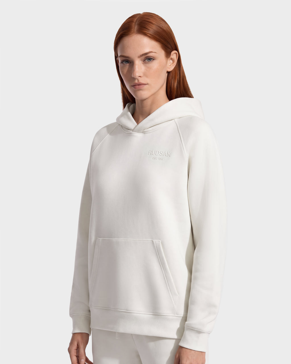 A person wearing a cream-colored Rudsak BRIAR - 9525001 Vanilla W unisex hoodie featuring a front pocket and embossed chest text for a casual look.