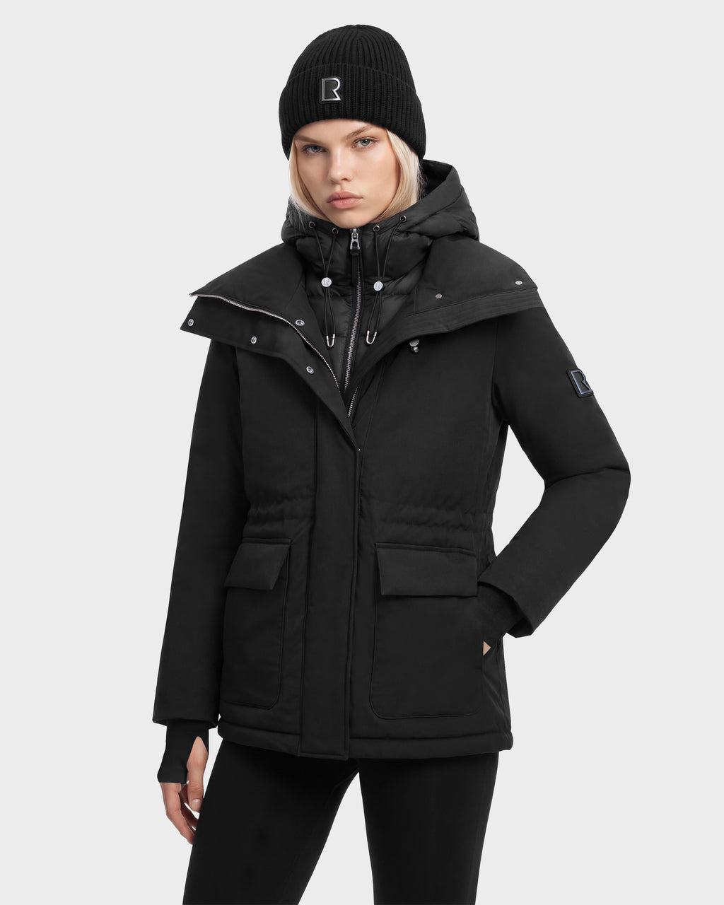 A person wears a black BHIMA - 8126515 Black hooded parka with a waist drawstring, large front pockets, and an internal zipped vest layer, paired with a matching black beanie.